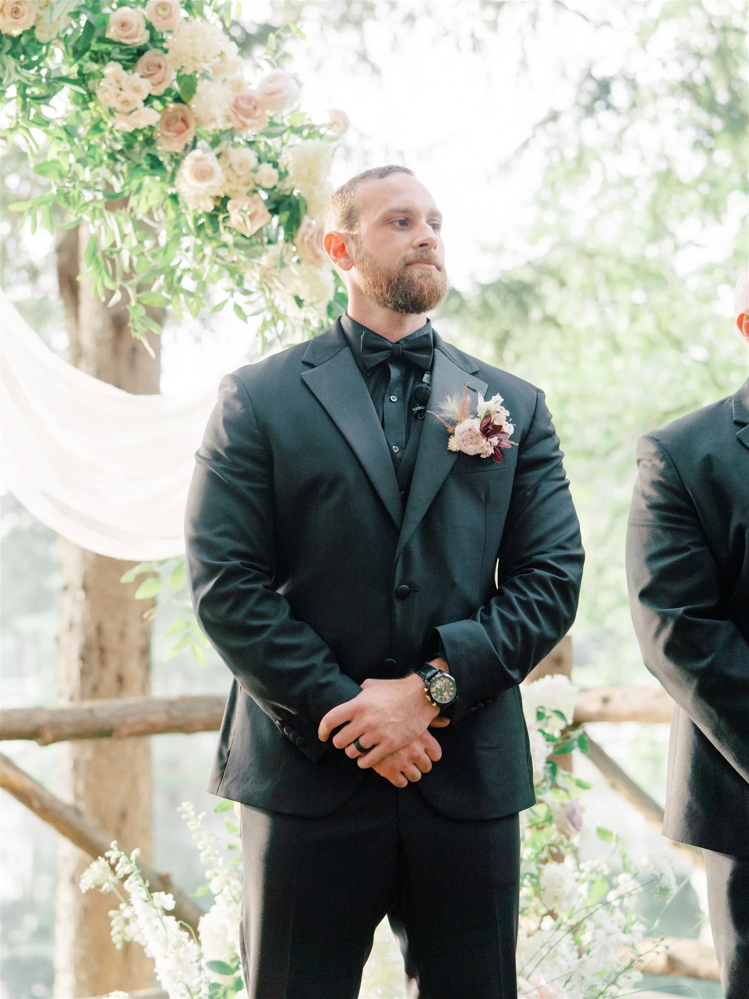 A man in a black tuxedo with a black bow tie and a floral boutonniere, standing outdoors during a wedding ceremony, with greenery and floral arrangements in the background.