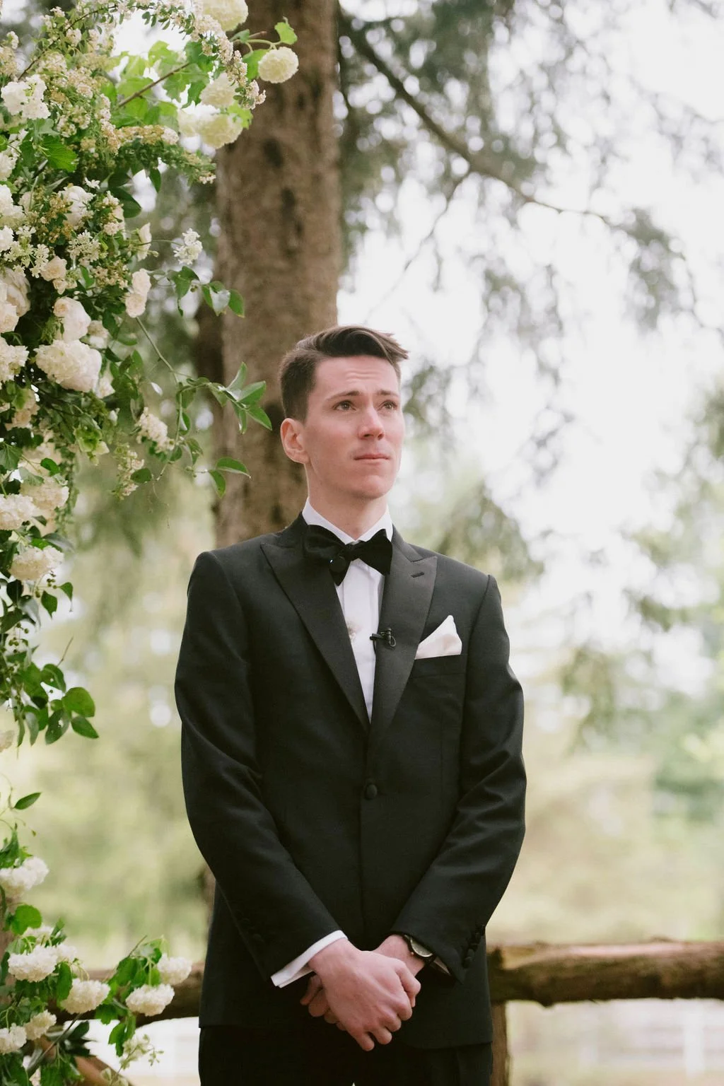 A young man in a black tuxedo with a bow tie and white pocket square standing outdoors near a tree and a floral decoration, with a somber expression on his face.
