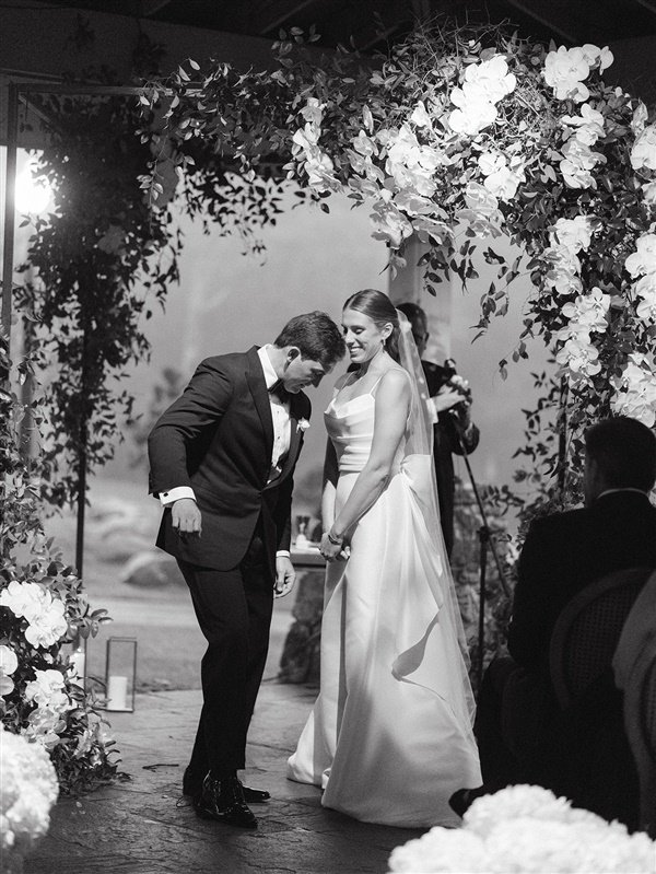 A black and white photograph of a couple at their wedding ceremony, standing under an arch decorated with flowers. The groom is bowing slightly, and the bride is smiling.