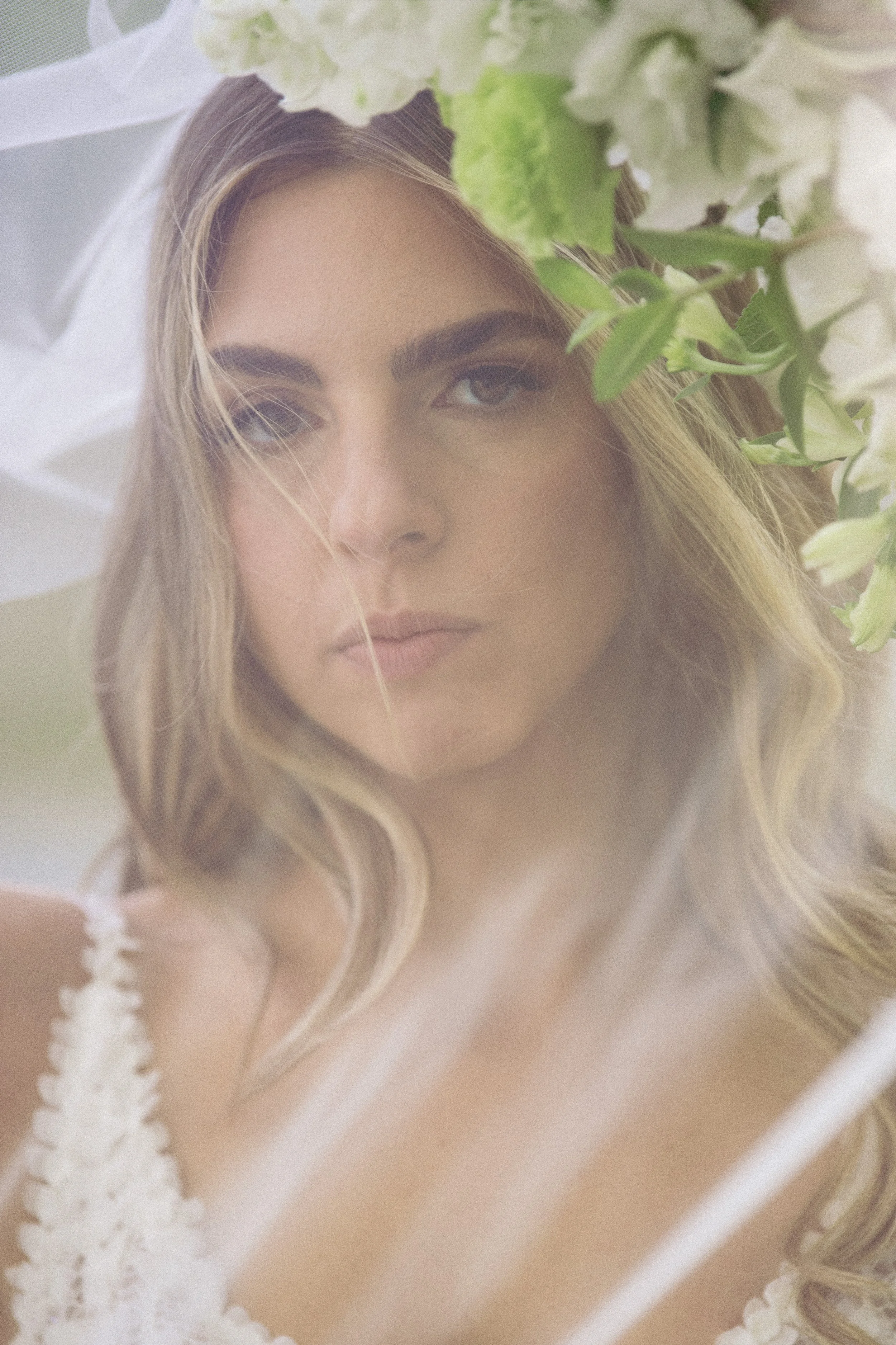 Close-up of a woman with long wavy hair, partially obscured by white and green flowers, with a soft focus and natural light.