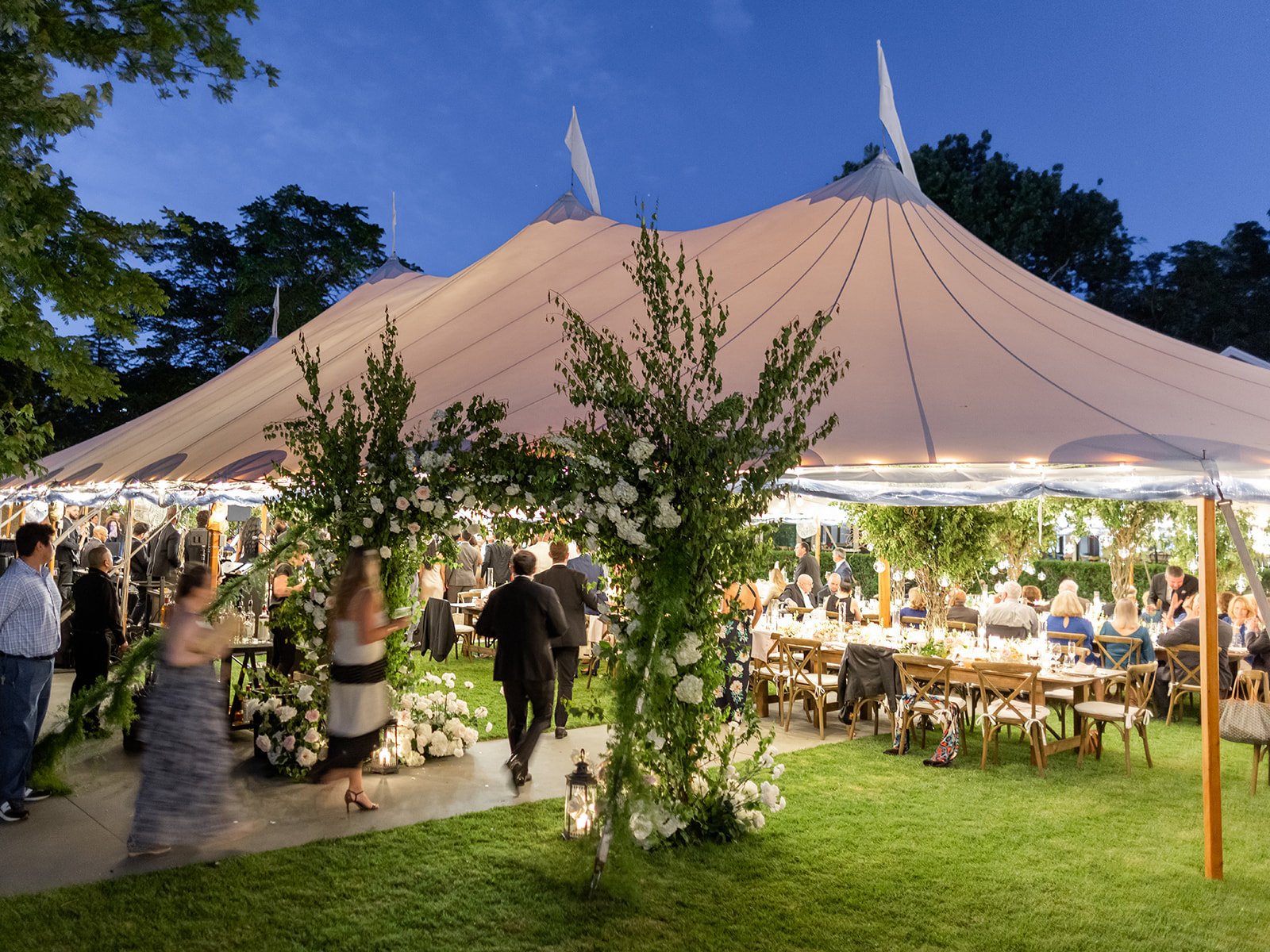 Outdoor wedding reception under a large white tent with string lights, floral arrangements, and guests seated at tables during evening.