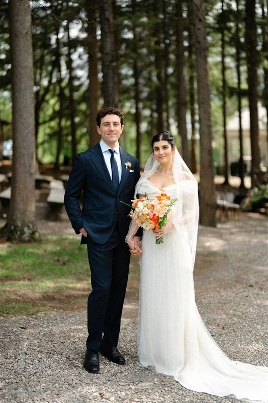 A bride and groom standing outdoors in a wooded area, holding hands, on their wedding day. The groom is in a navy suit and tie, and the bride is in a white wedding dress with a veil, holding a bouquet of flowers.
