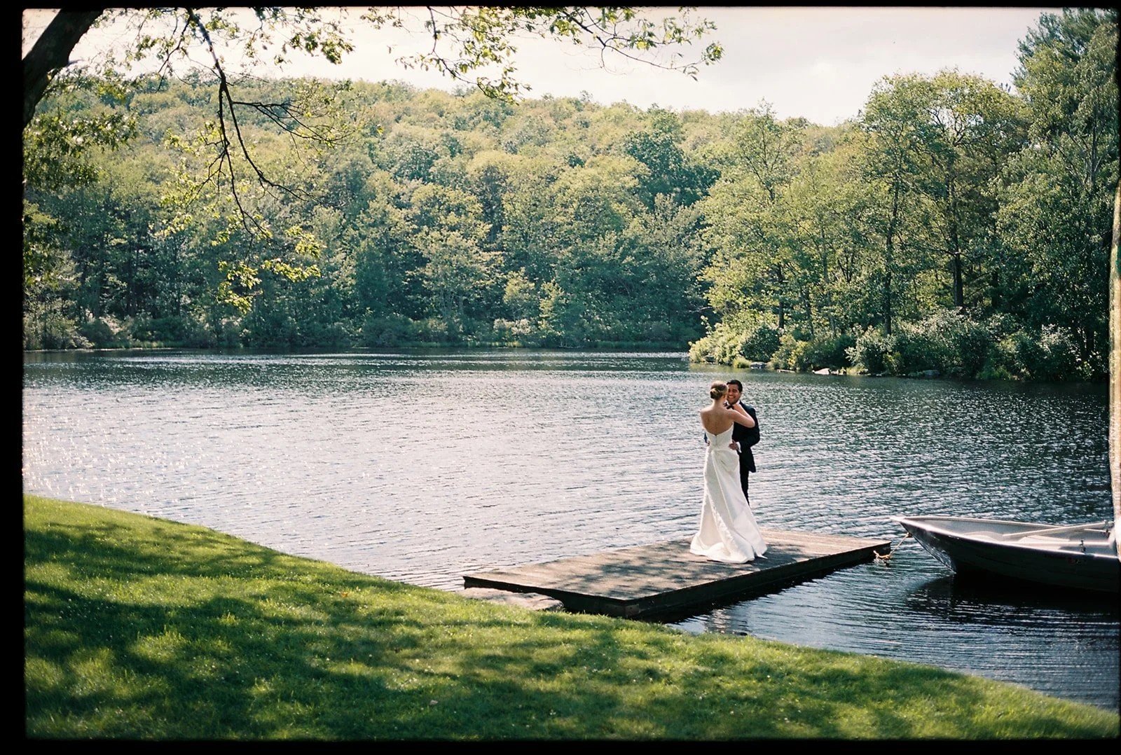 A couple in wedding attire dancing on a dock by a lake surrounded by trees.