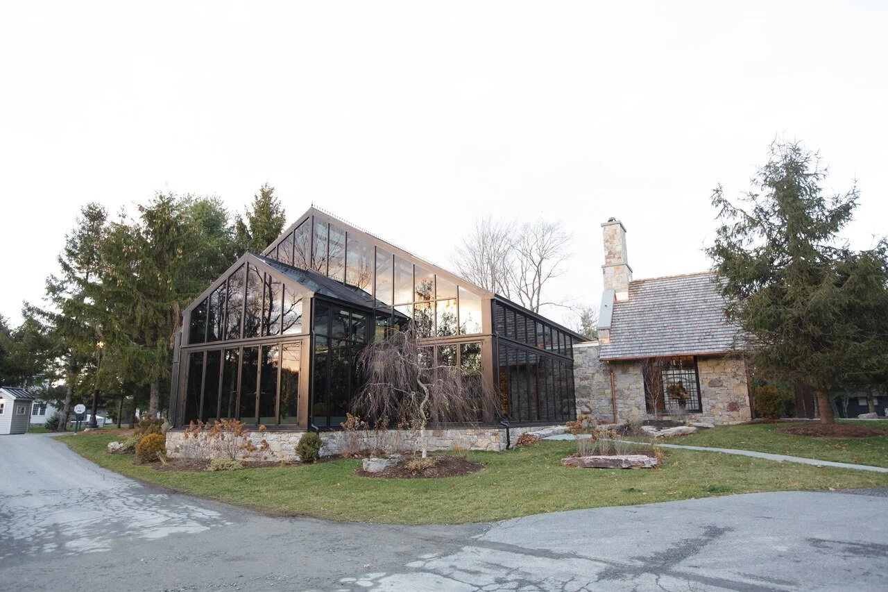 A house with a traditional stone exterior and a chimney, attached to a modern glass extension, surrounded by trees and a driveway.