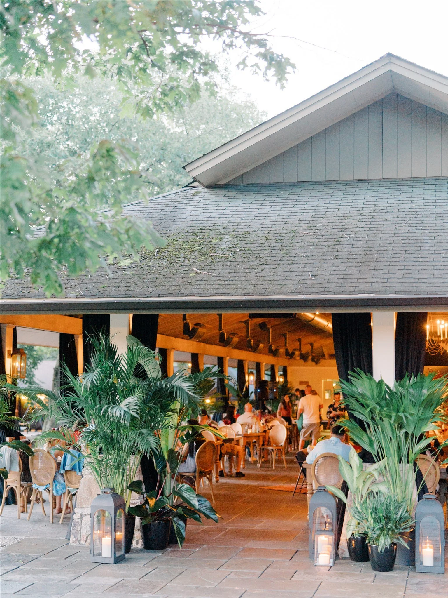 An outdoor patio of a restaurant or event space during the evening with large potted plants, lanterns with candles, and people dining inside