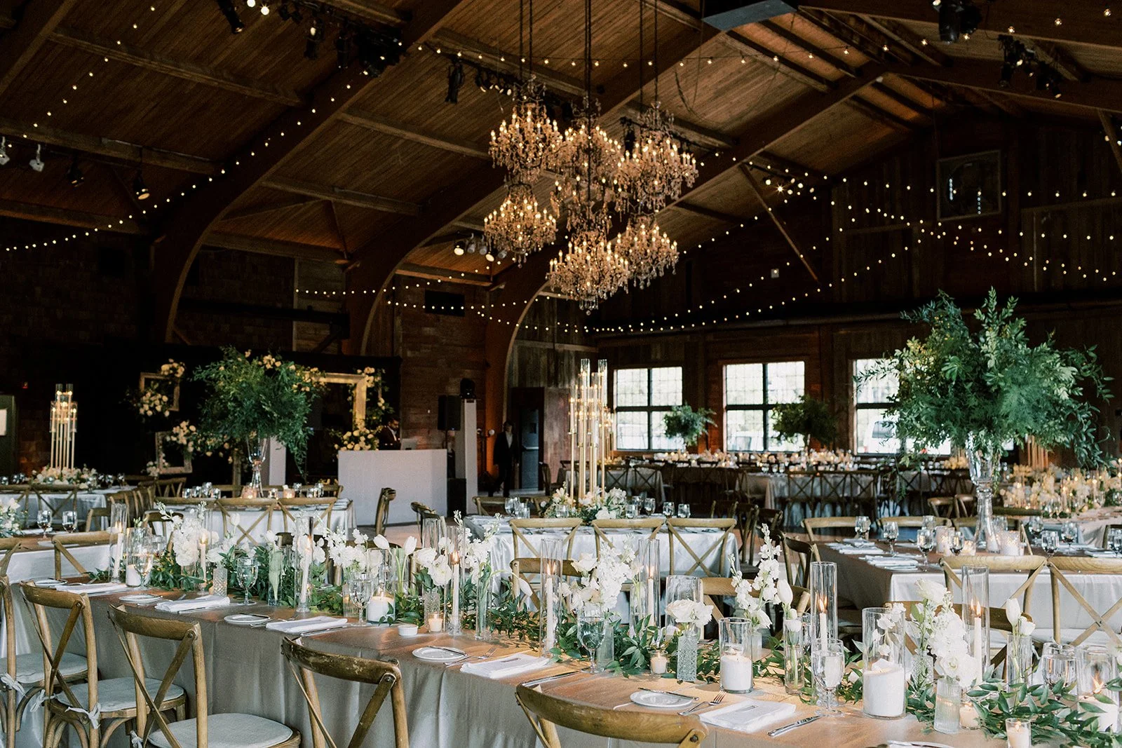 Elegant indoor wedding reception decorated with white flowers, candles, and greenery, with large chandeliers hanging from a wooden ceiling.