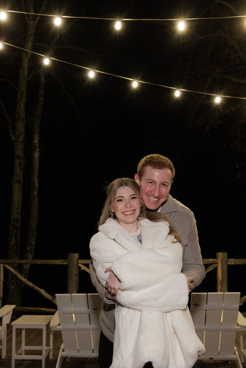 A smiling young woman wrapped in a white fur coat is hugging a smiling young man at night outdoors, with string lights overhead and a wooden railing and benches in the background.