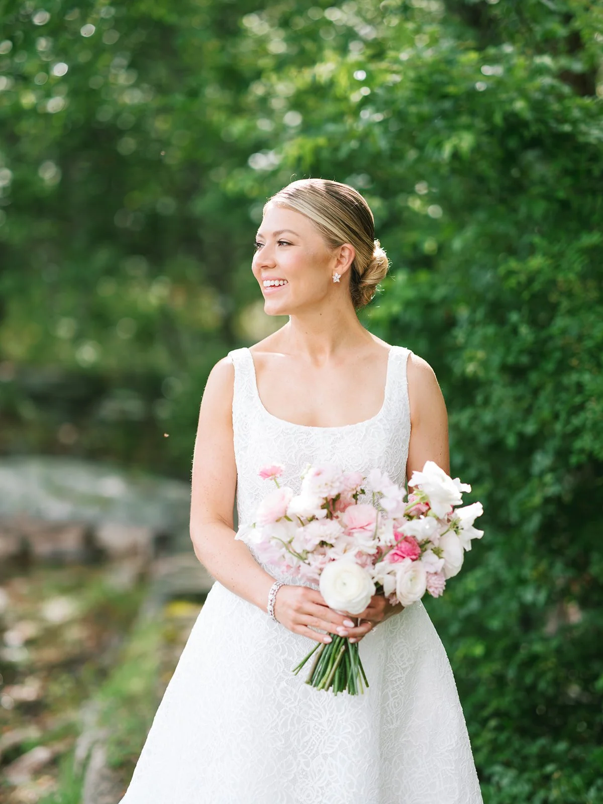 A bride in a white lace wedding dress holding a bouquet of pink and white flowers outdoors, smiling and looking to her left with a background of green trees.