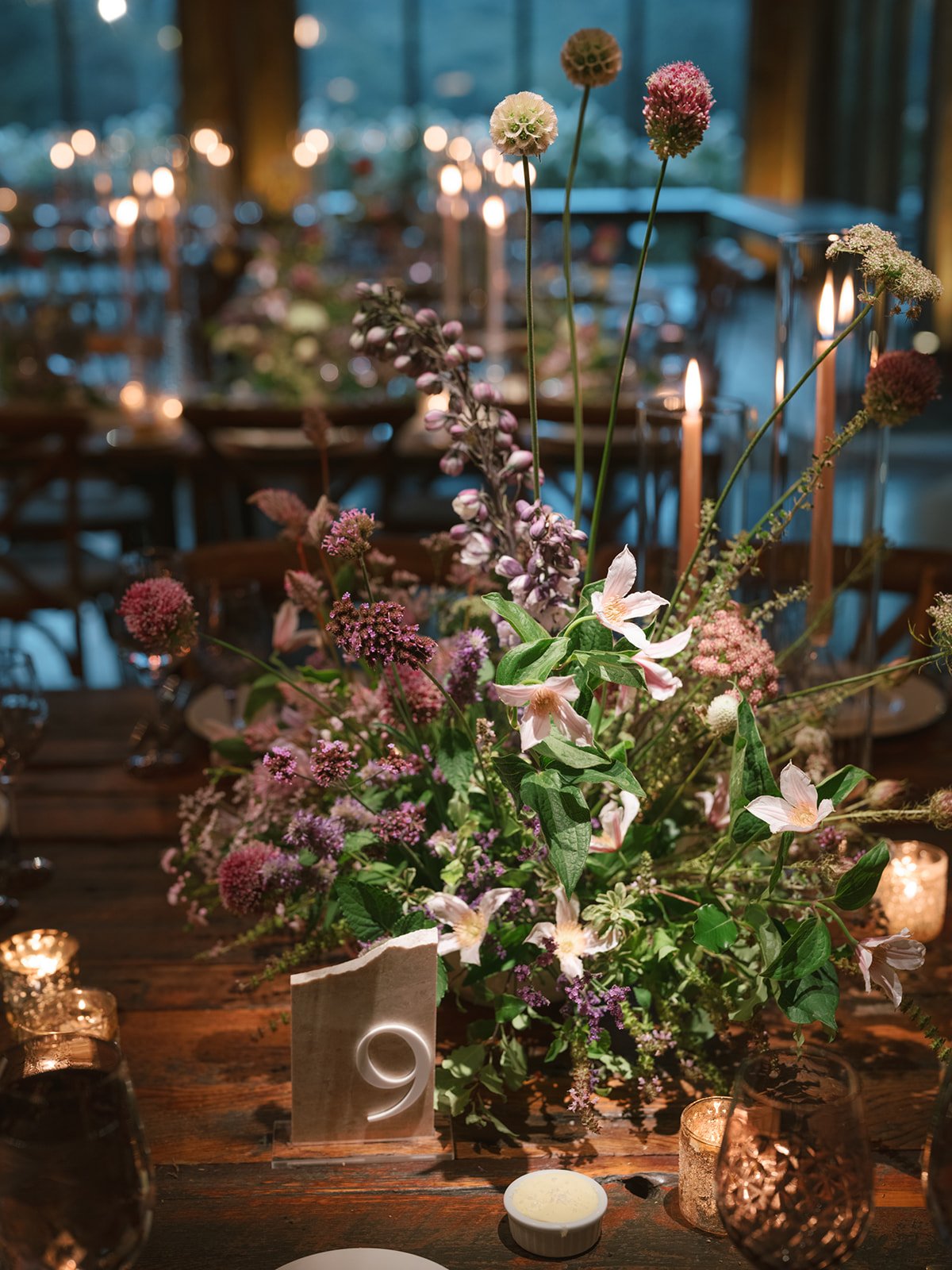 Floral centerpiece with pink and purple flowers on a wooden table, surrounded by candles and a table number '9', in a dimly lit event space.