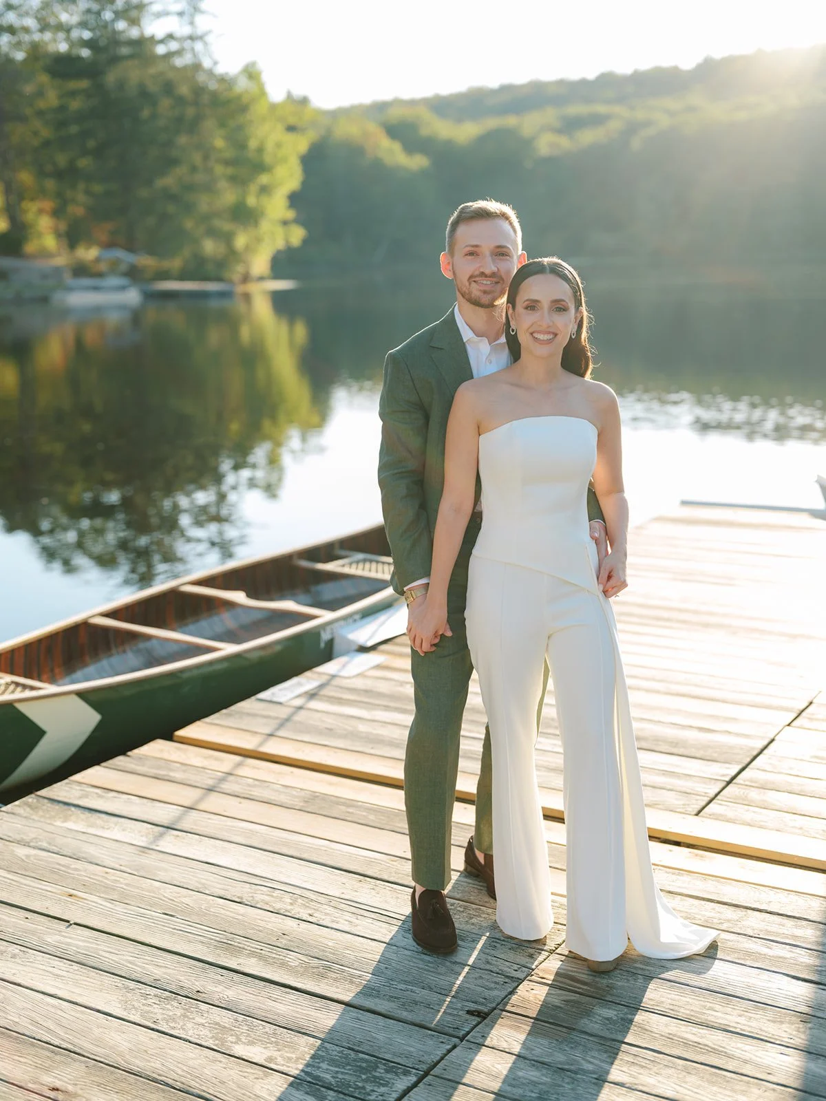 A couple dressed in wedding attire standing on a wooden dock near a lake, holding hands, with boats and trees in the background.
