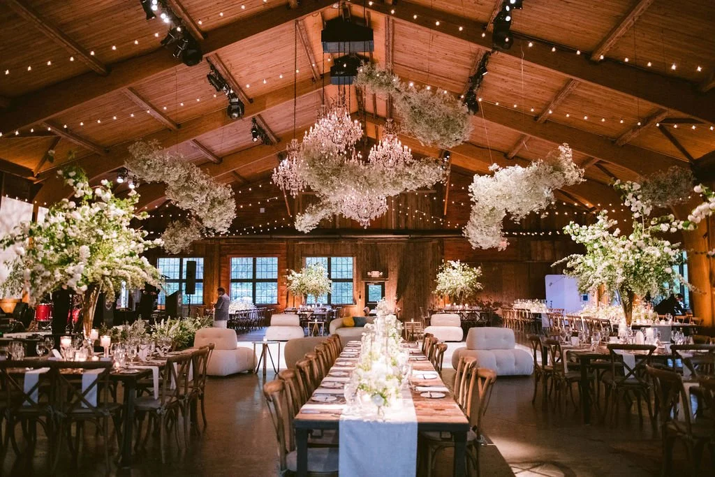 Beautifully decorated rustic wedding reception hall with wooden ceiling, large floral centerpieces, and warm lighting.
