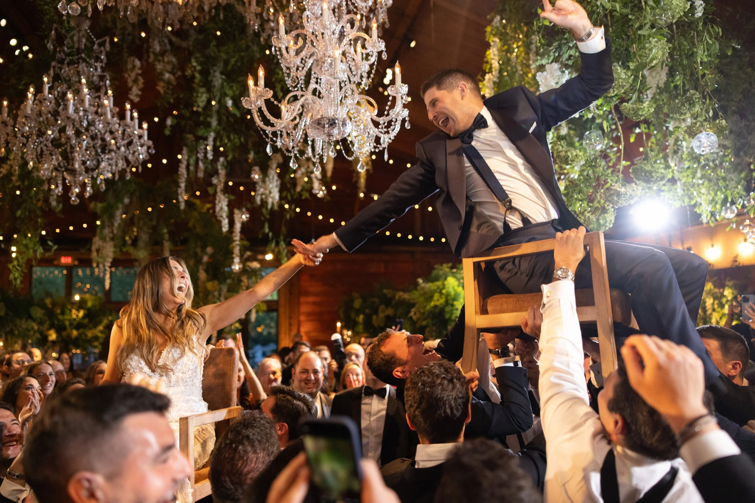 A man in a tuxedo sits on a chair being lifted above a crowd during a celebration, reaching out and holding hands with a smiling woman in a white dress. The scene is decorated with chandeliers and greenery.