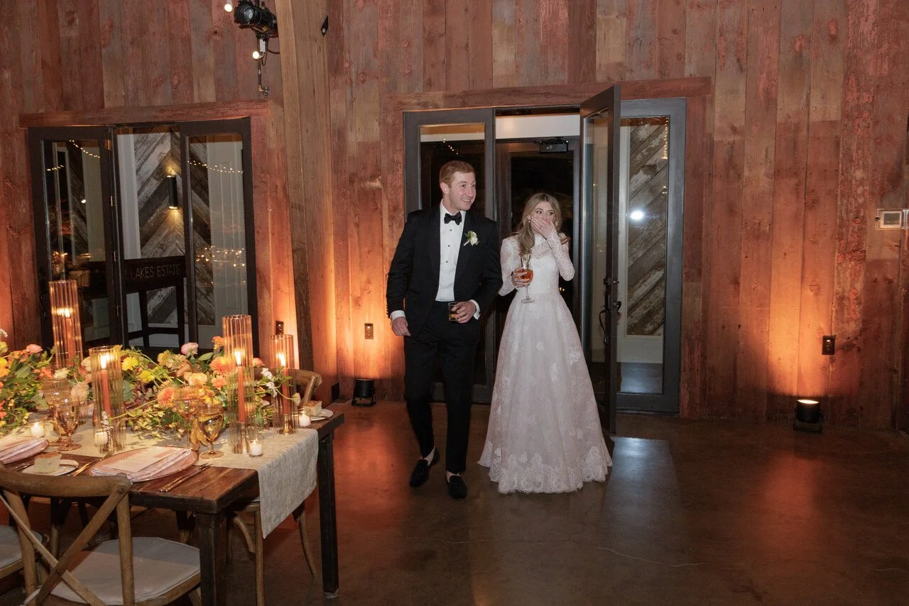 Bride and groom entering a reception hall with rustic wooden walls, decorated with candles and floral arrangements, at a wedding celebration.