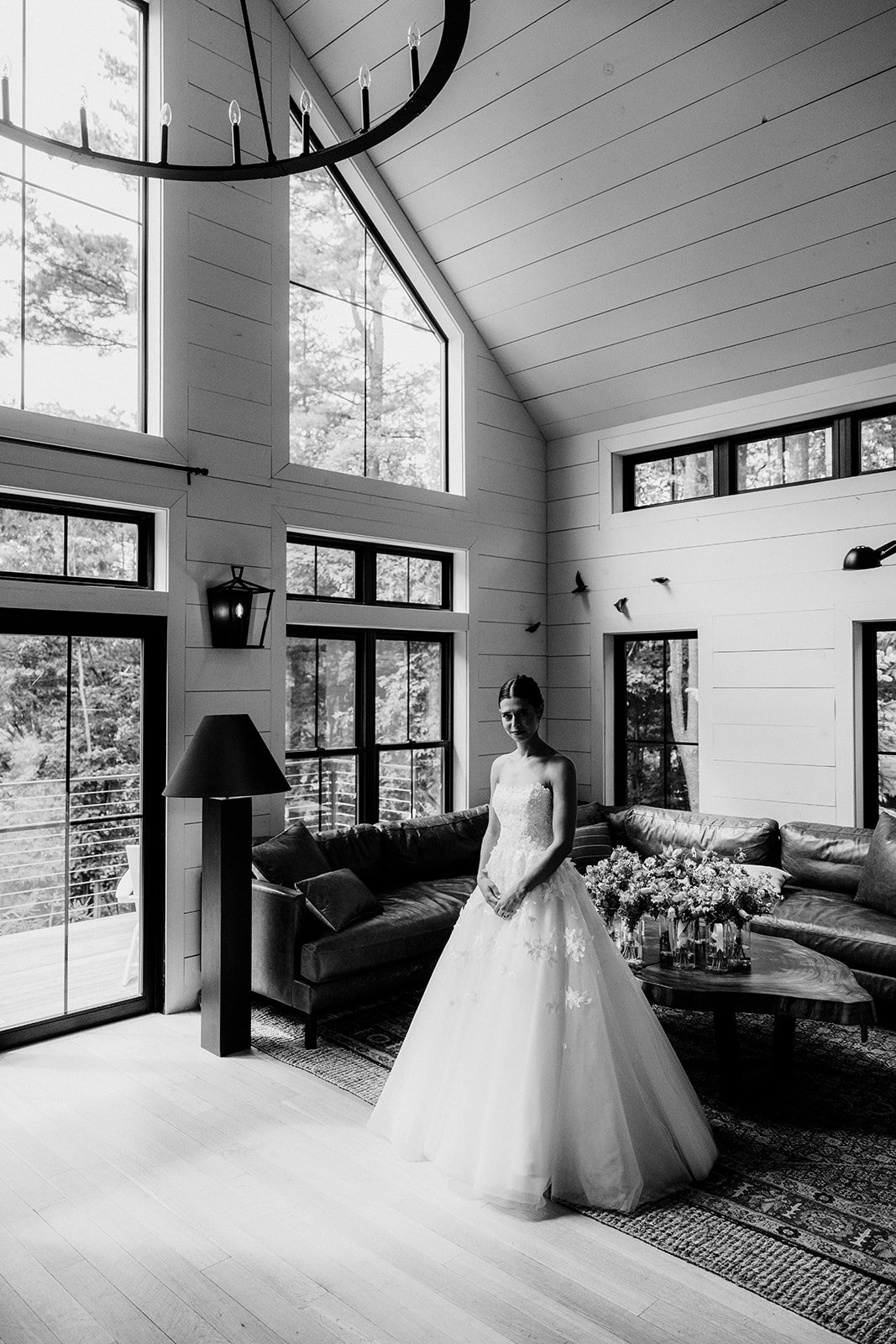 A bride in a wedding dress standing inside a modern living room with large windows, a sofa, a coffee table with flowers, and natural light from outside.