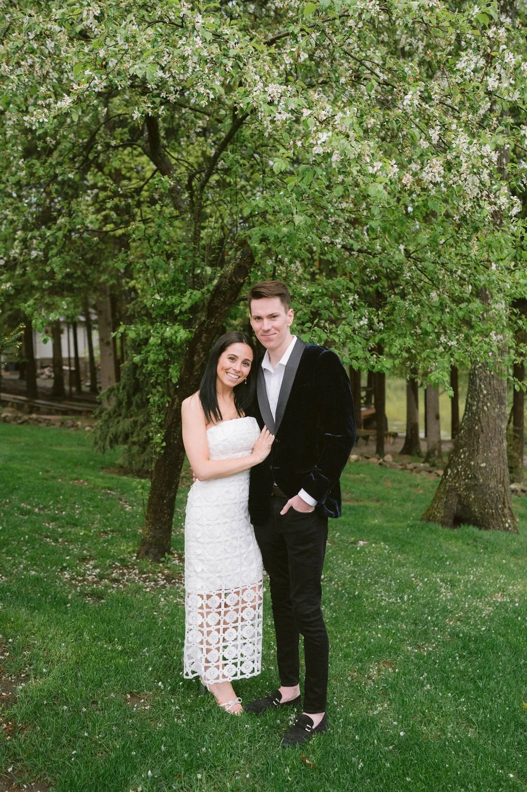 A smiling couple stands together outdoors under blooming trees, the woman in a white lace dress and the man in a black suit jacket and dress pants.