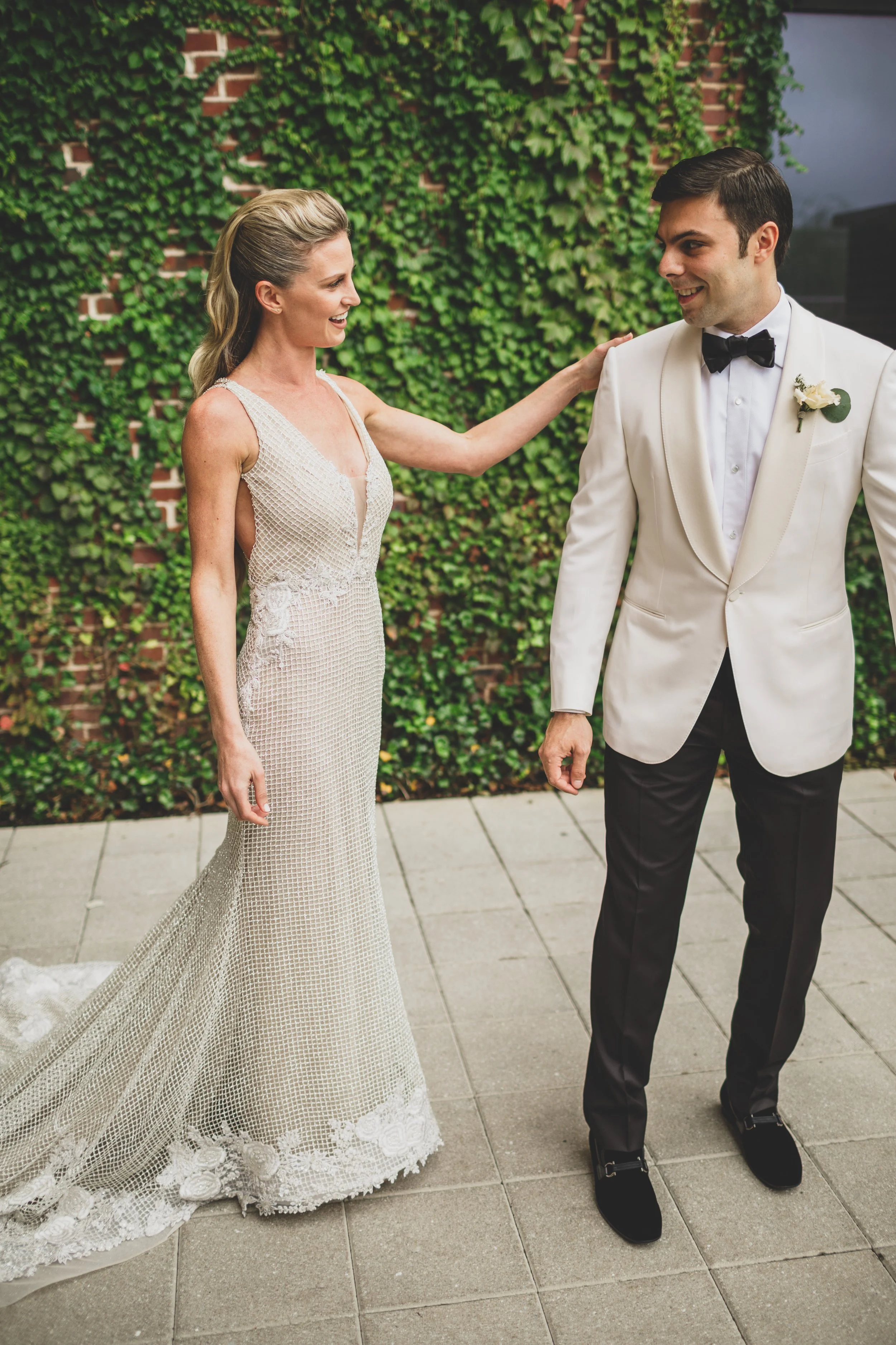 A bride in a long, white, patterned dress with lace details is touching the shoulder of a groom in a white tuxedo jacket, black pants, and a black bow tie, standing outside against a wall covered with green ivy.