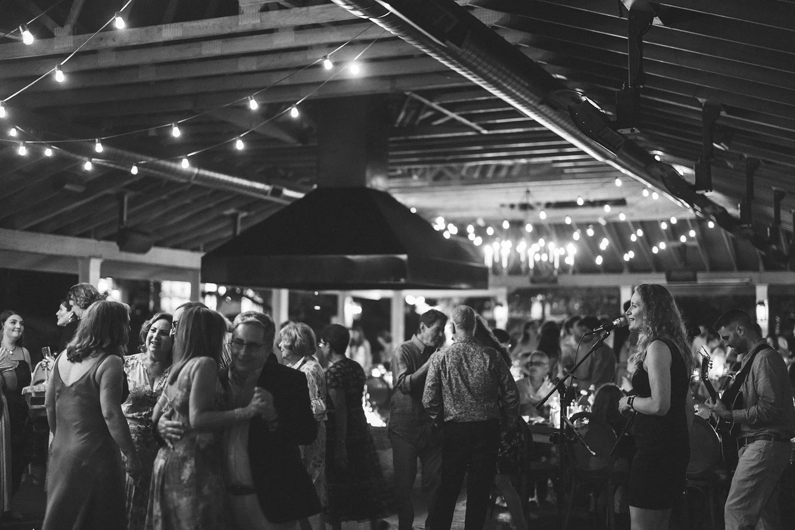 People enjoying a live music performance at an indoor event space with string lights and a high, open ceiling.