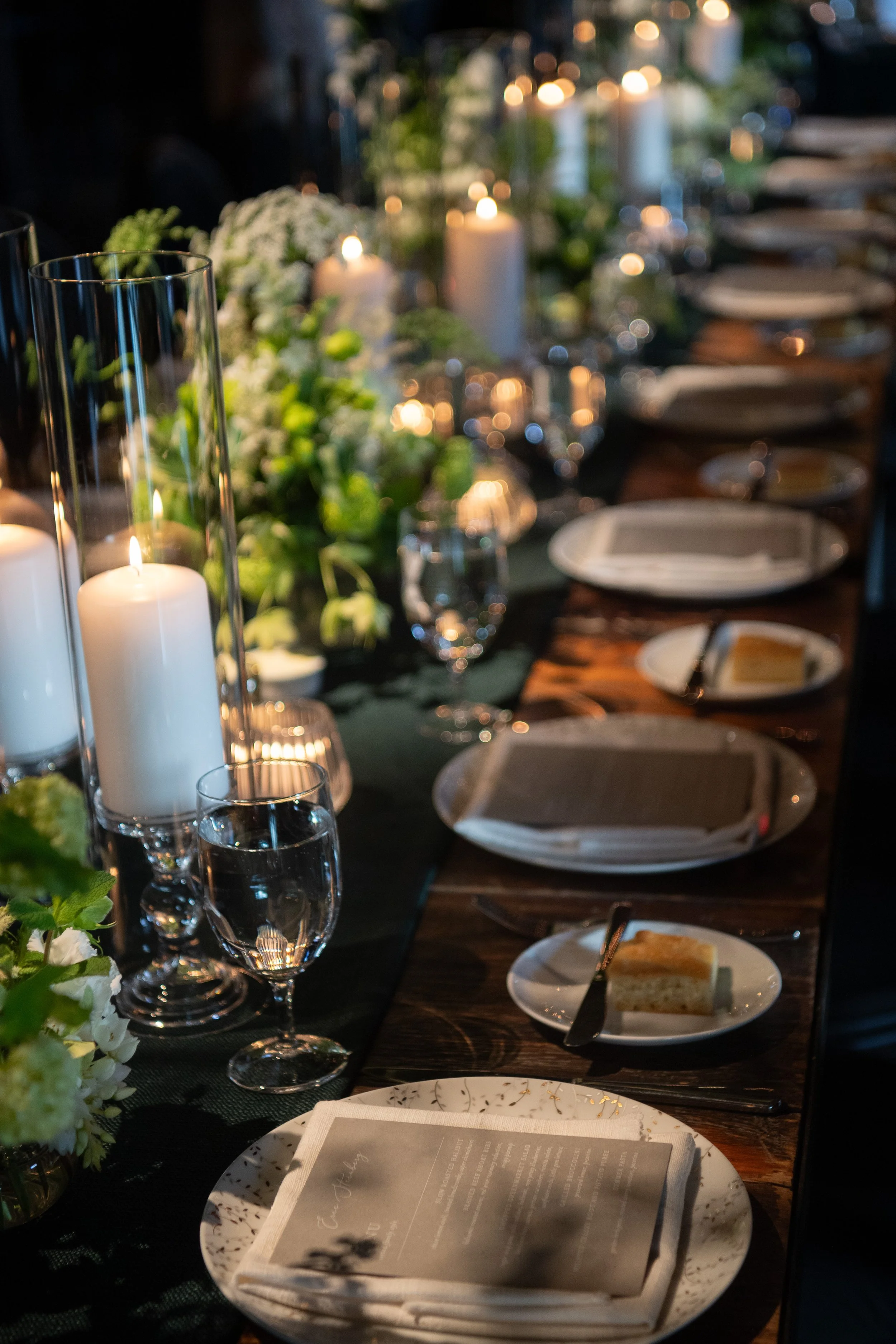 A decorated dining table set for a meal with plates, utensils, wine glasses, and candles. There are flowers and greenery as centerpieces, with a warm, elegant ambiance.