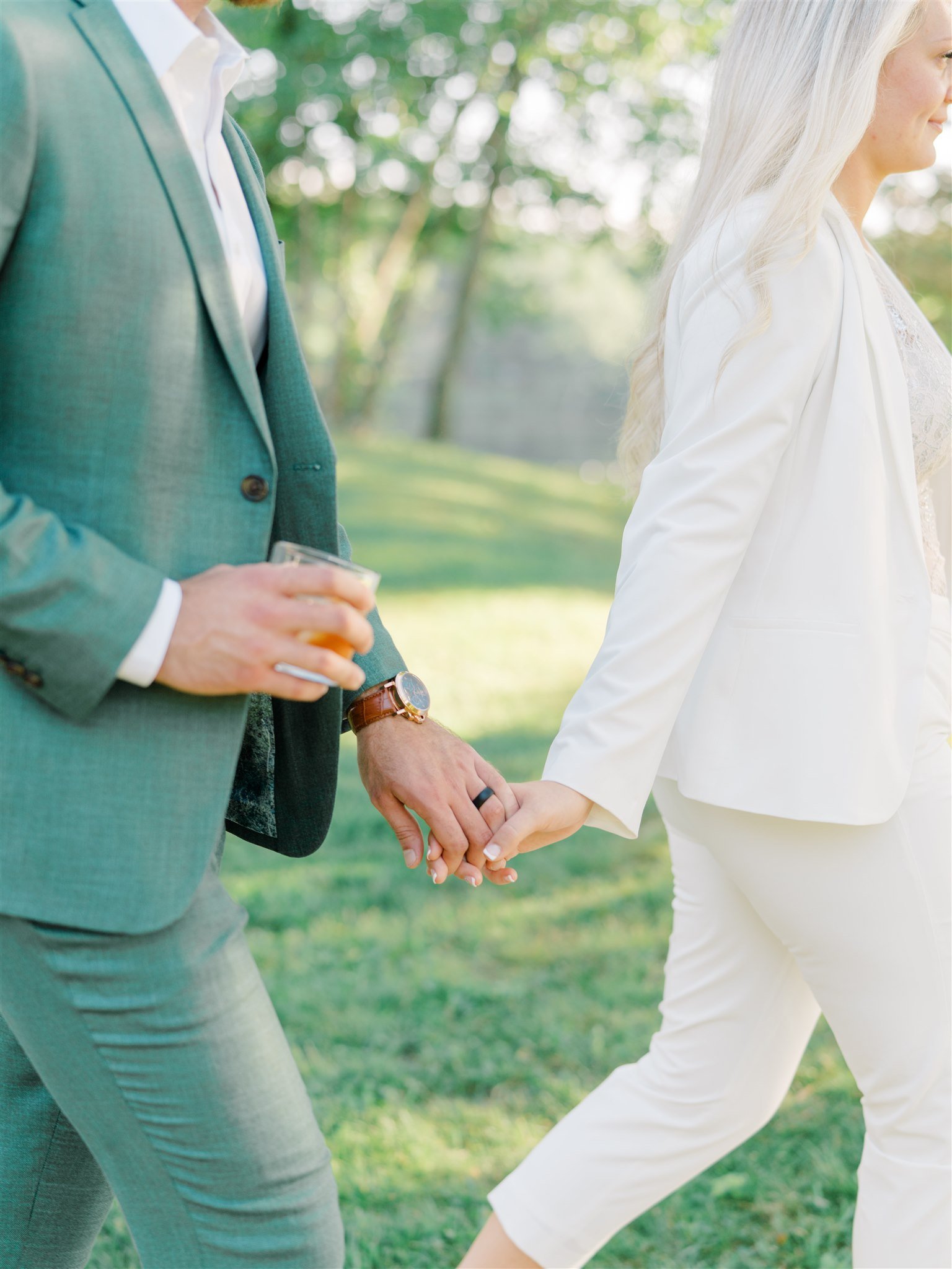 Close-up of a man and woman holding hands outdoors during daytime, woman with white blazer and pants, man in green suit holding a drink.