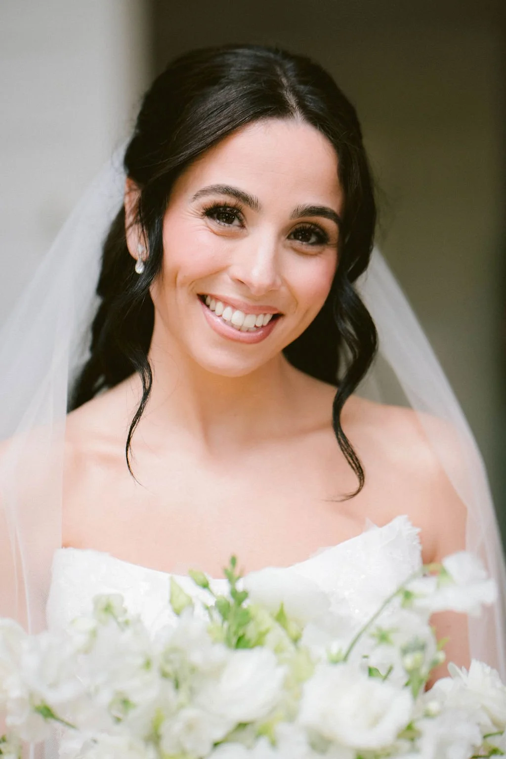 Close-up portrait of a smiling bride with dark hair, wearing a white wedding dress and veil, holding a bouquet of white flowers.