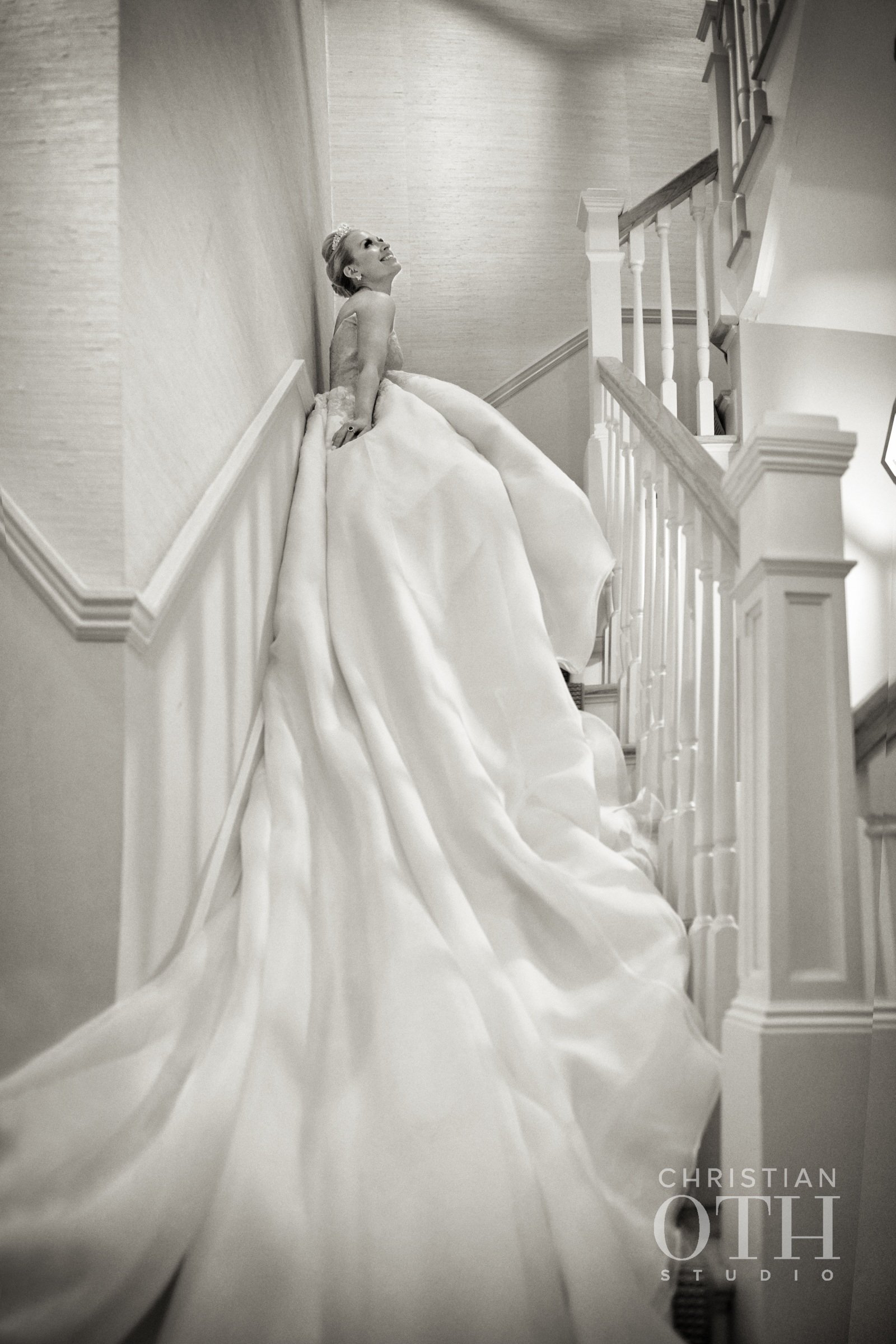 A bride in a wedding gown standing on a staircase, looking up and smiling.