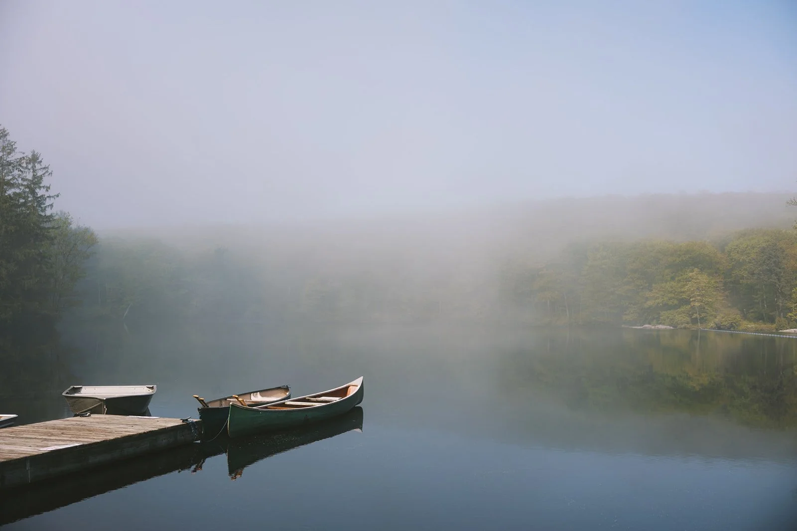 A foggy lake with three small boats tied to a wooden dock, with trees in the background