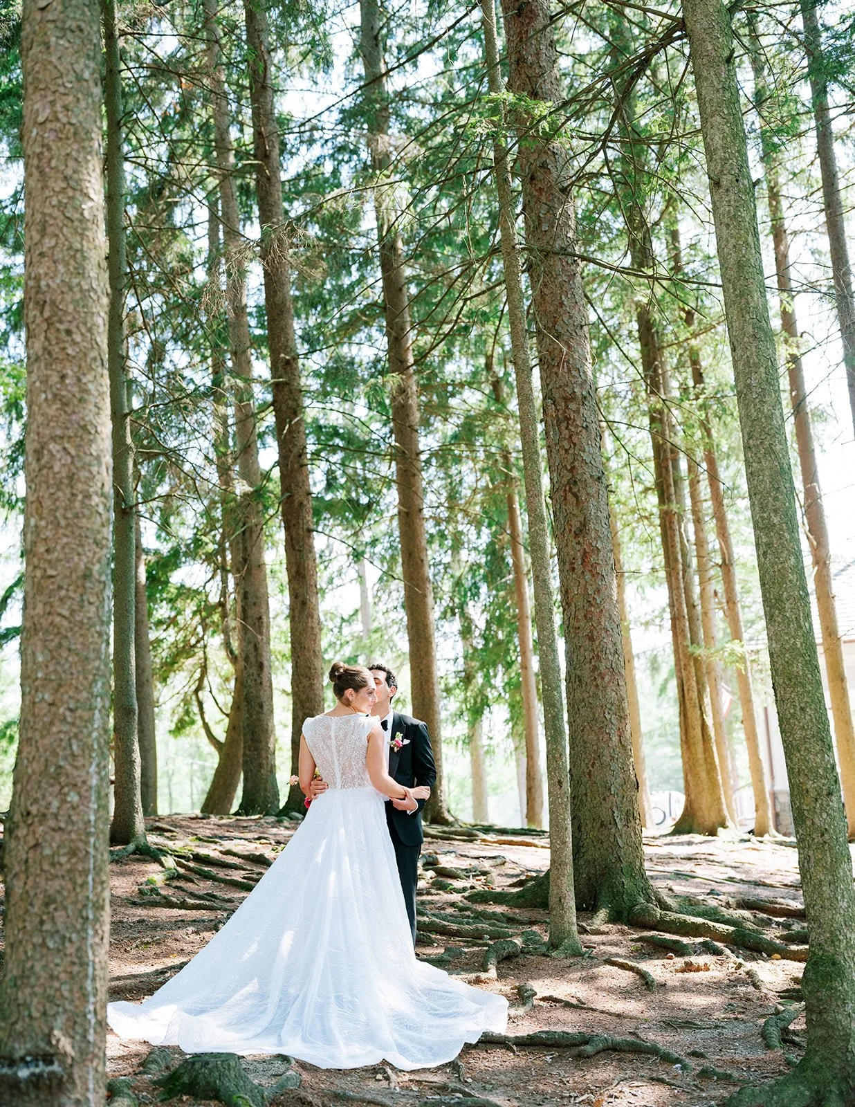 A bride and groom in wedding attire standing in a forest with tall trees, holding hands and gazing at each other.