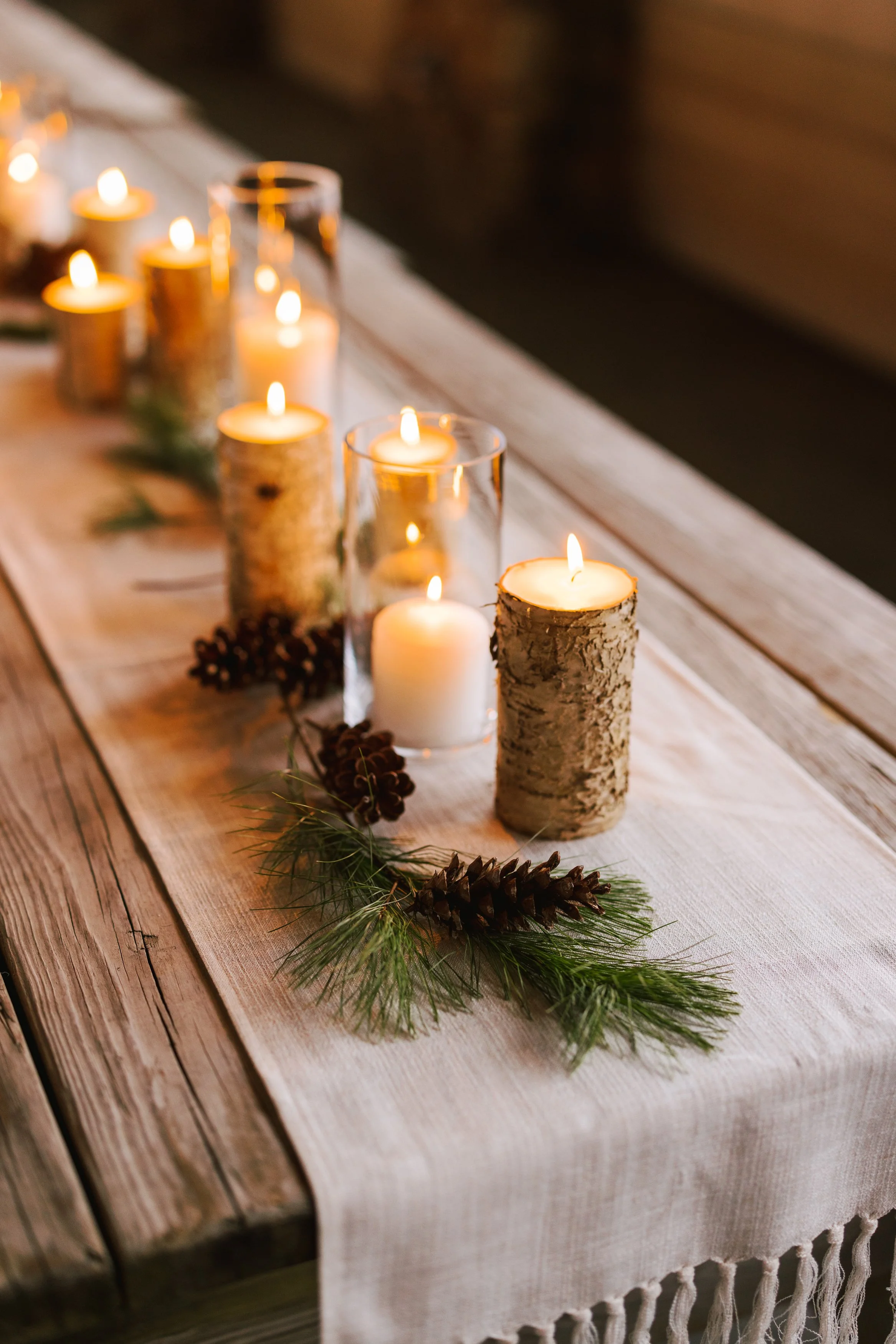 Table decorated with candles in glass holders, pinecones, and pine branches, set on a rustic wooden table with a white table runner.
