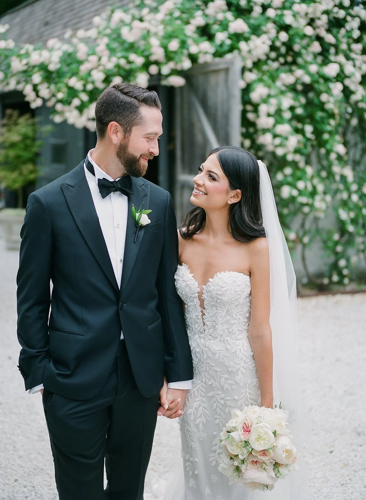 A bride and groom standing outdoors, holding hands and smiling at each other, with blooming flowers and a rustic building in the background.
