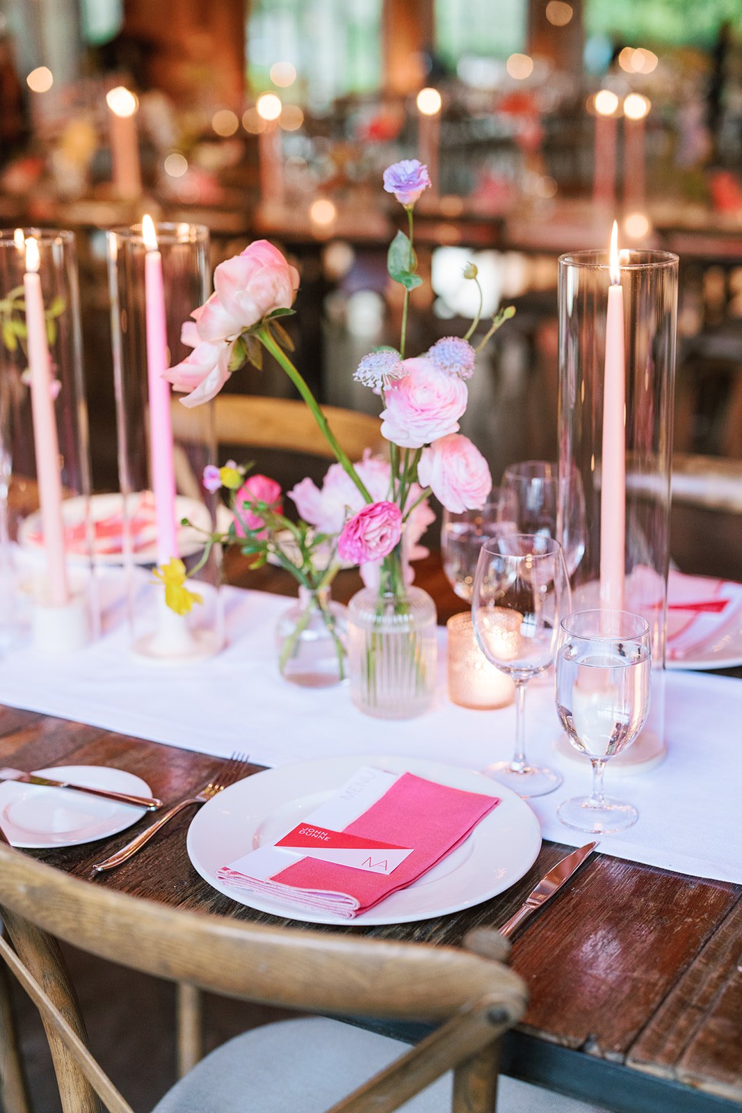 Decorated dining table with pink flowers in a glass vase, lit candles in glass holders, pink napkin on a white plate, wine glasses, and cutlery, set in a warmly lit rustic venue.