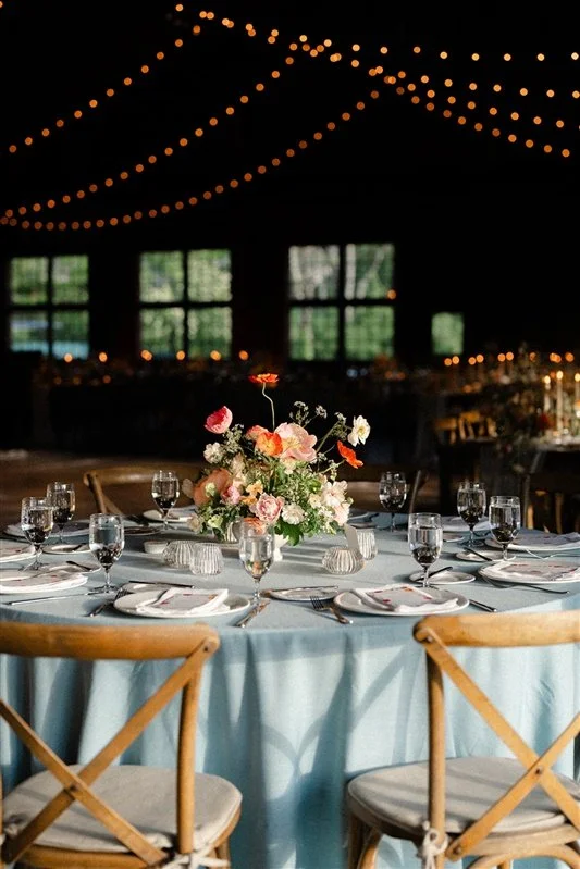 A wedding reception table decorated with a floral centerpiece, wine glasses, and place settings, under string lights in a dimly lit venue with large windows.