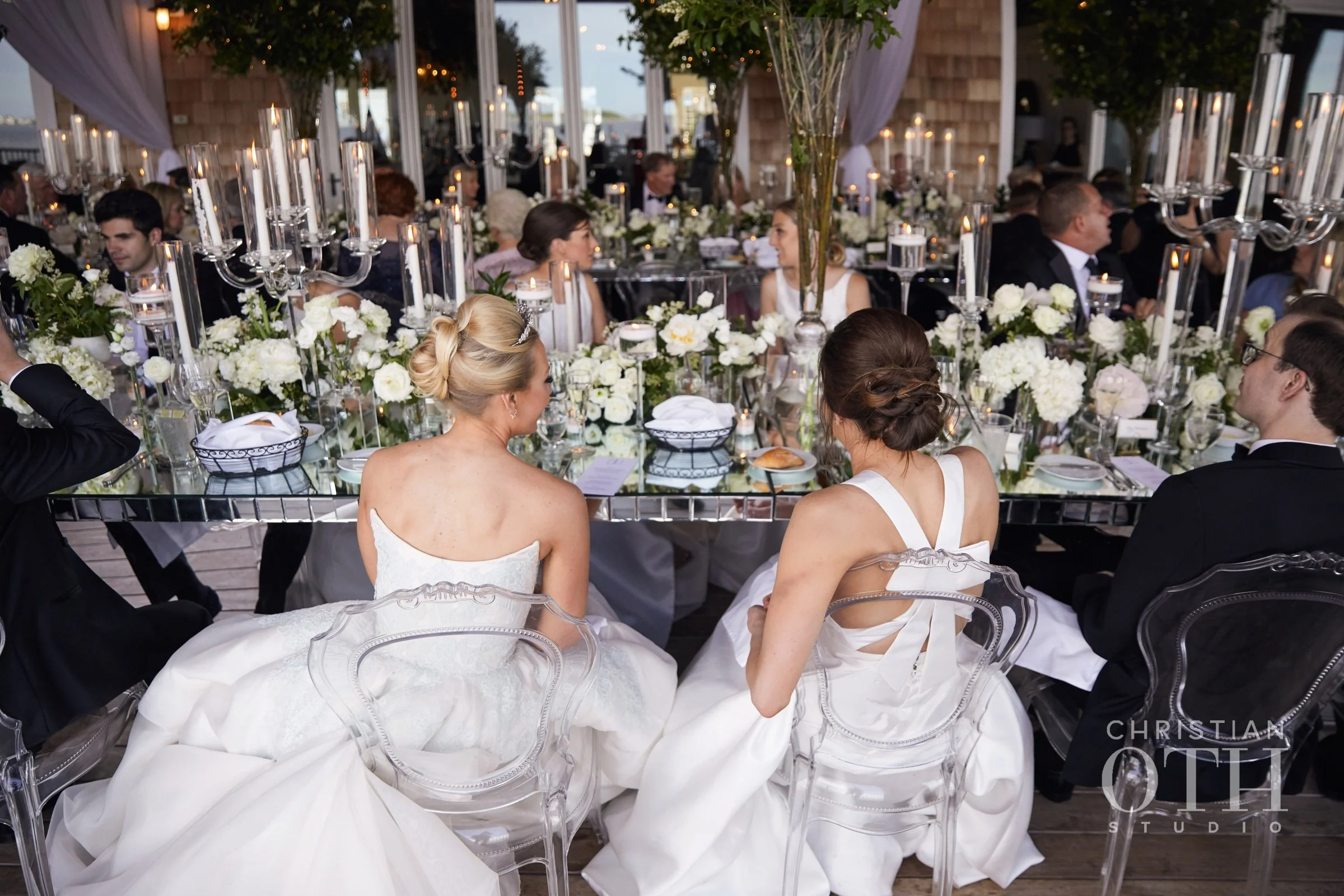 Elegant wedding reception with guests seated at a long table decorated with white flowers, candles, and tall candelabras, with brides in white gowns and grooms in black tuxedos visible from behind.