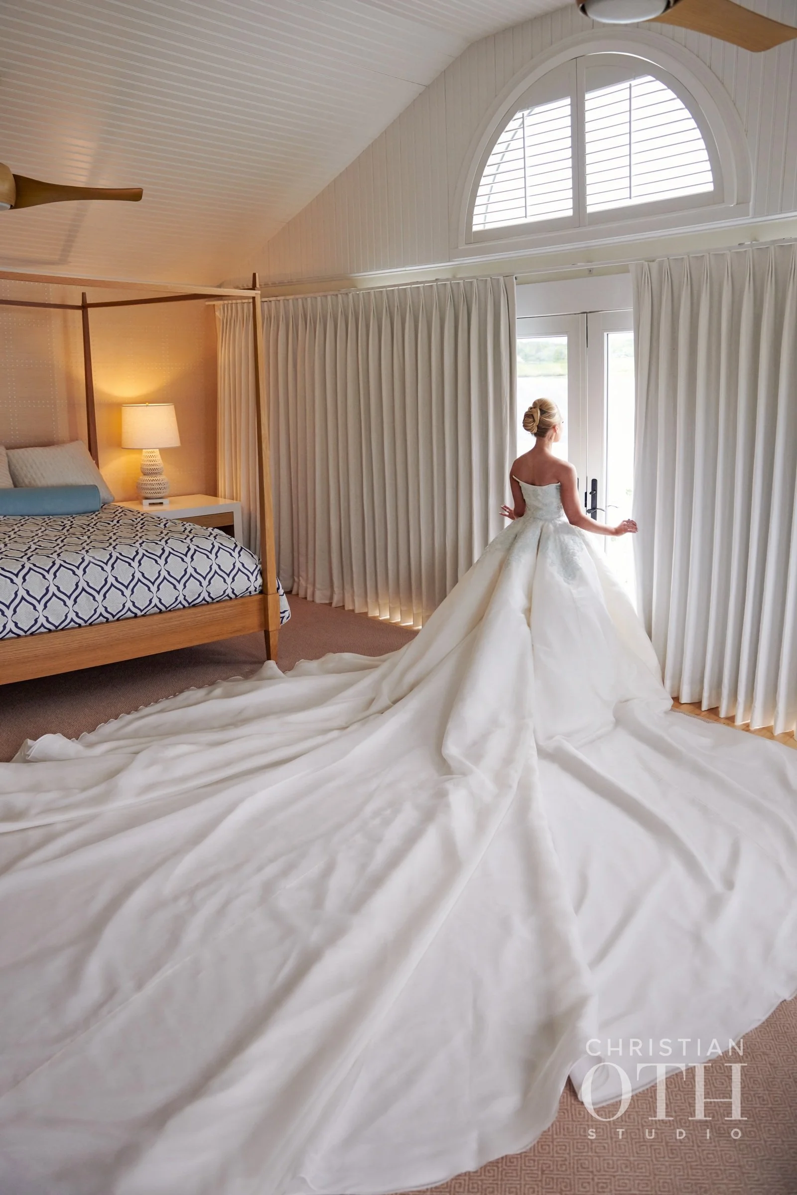 A bride in a white wedding gown standing by a window in a bedroom, with long train flowing on the floor.