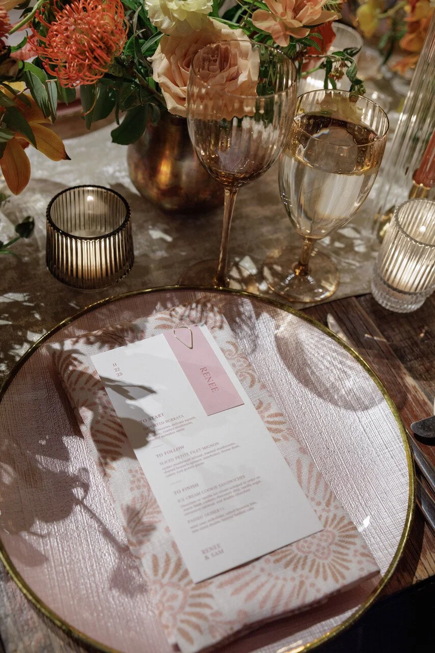 Elegant table setting with a menu on a pink patterned napkin, two glasses of water and wine, a floral centerpiece, and candle holders.