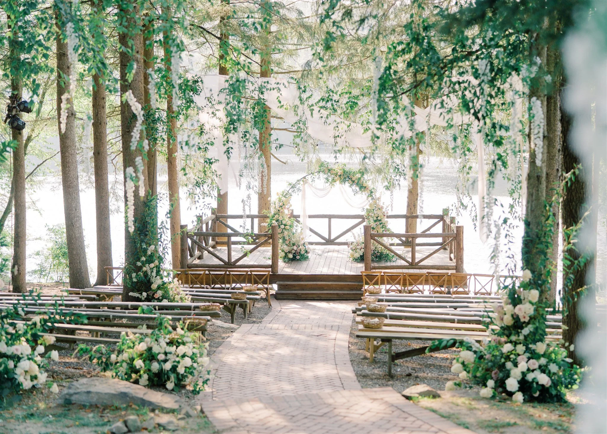 Outdoor wedding ceremony setup in a wooded area by a lake, with wooden aisle, flower arrangements, and a decorated arch at the altar.