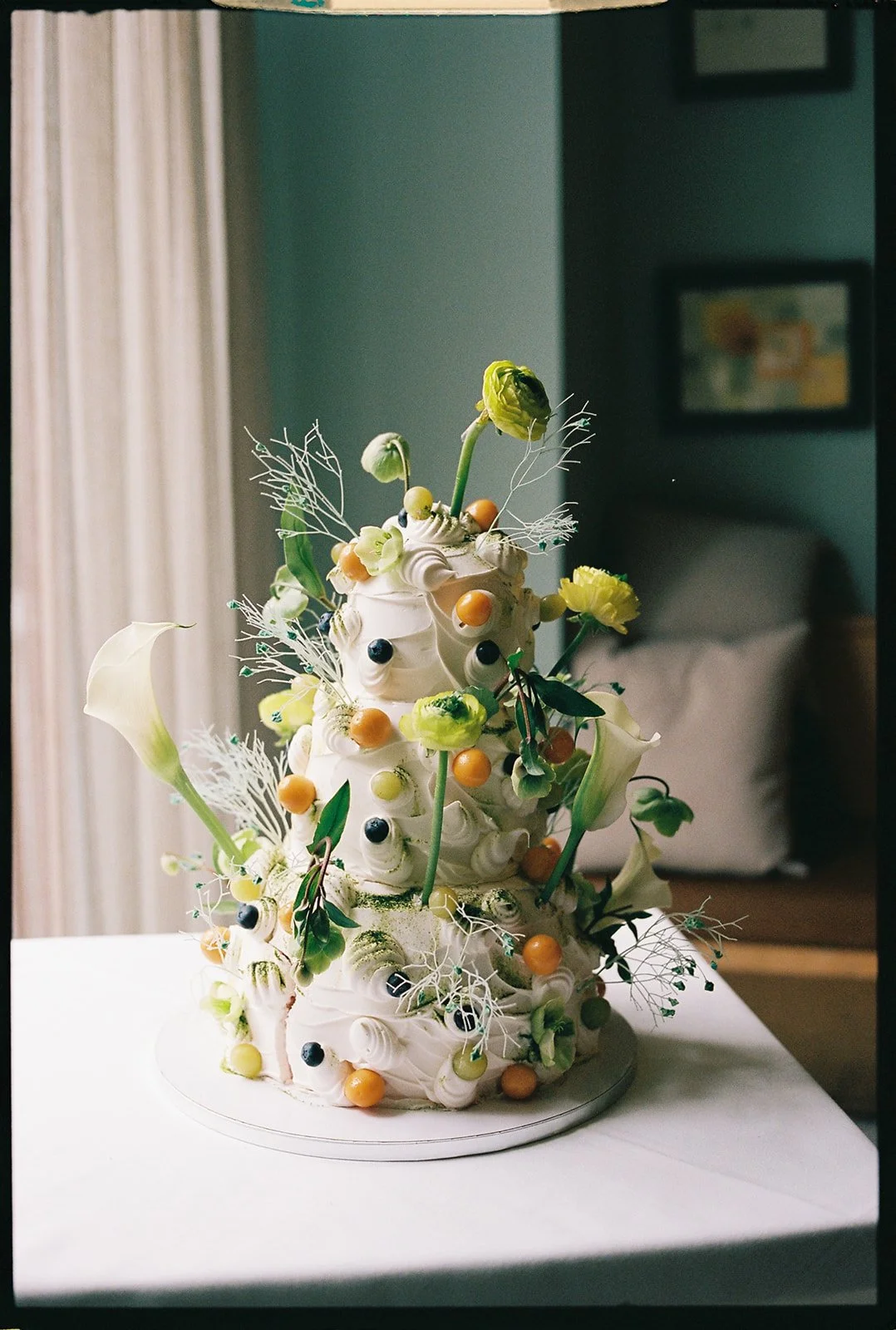 A sculptural cake decorated with white frosting, yellow and black berries, fresh flowers, and greenery, set on a table in a room with a bed and framed artwork in the background.