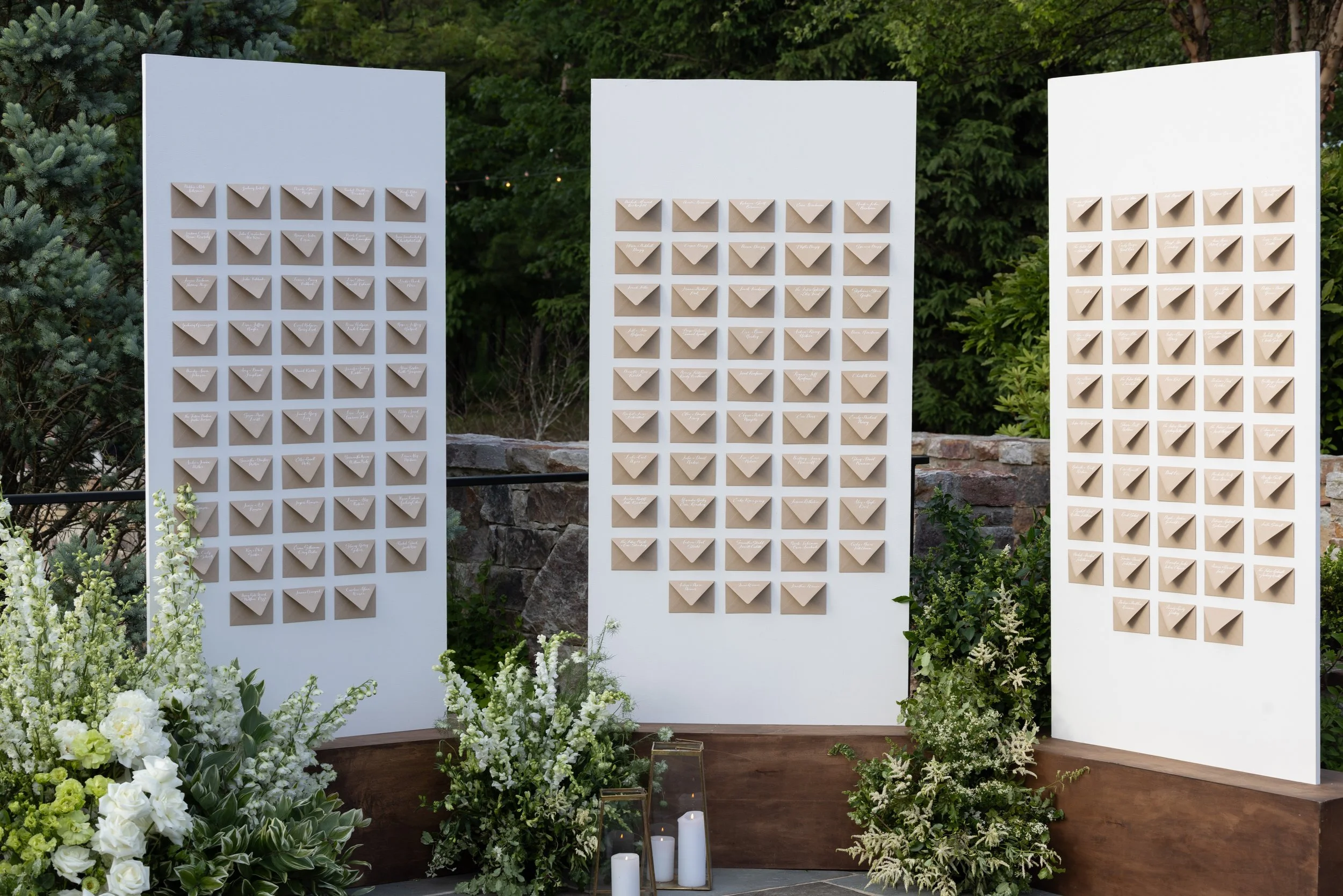 Three white display boards arranged outdoors, each with numerous small beige paper envelopes attached in neat rows, surrounded by green foliage and white flowers.