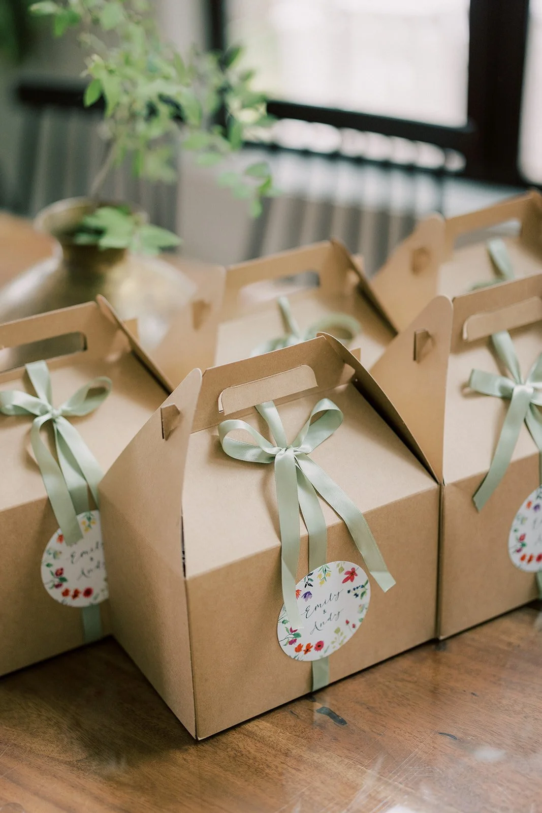Multiple brown gift boxes with light green ribbons and floral tags on a wooden table, with a blurred background of a window and a potted plant.