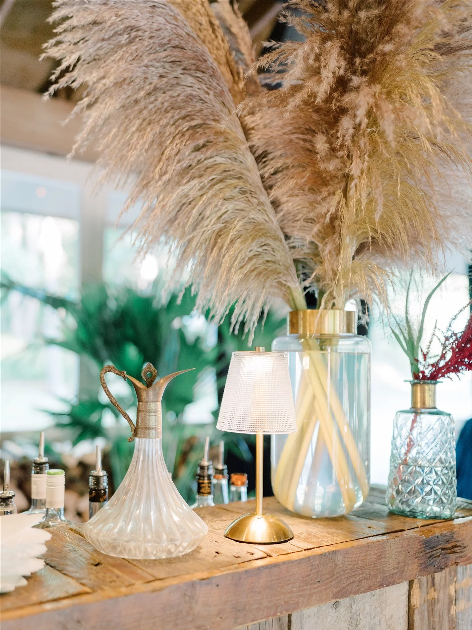 Decorative dried pampas grass in gold and glass vases on a rustic wooden table, accompanied by a small table lamp and various glass bottles or containers, in a well-lit room.