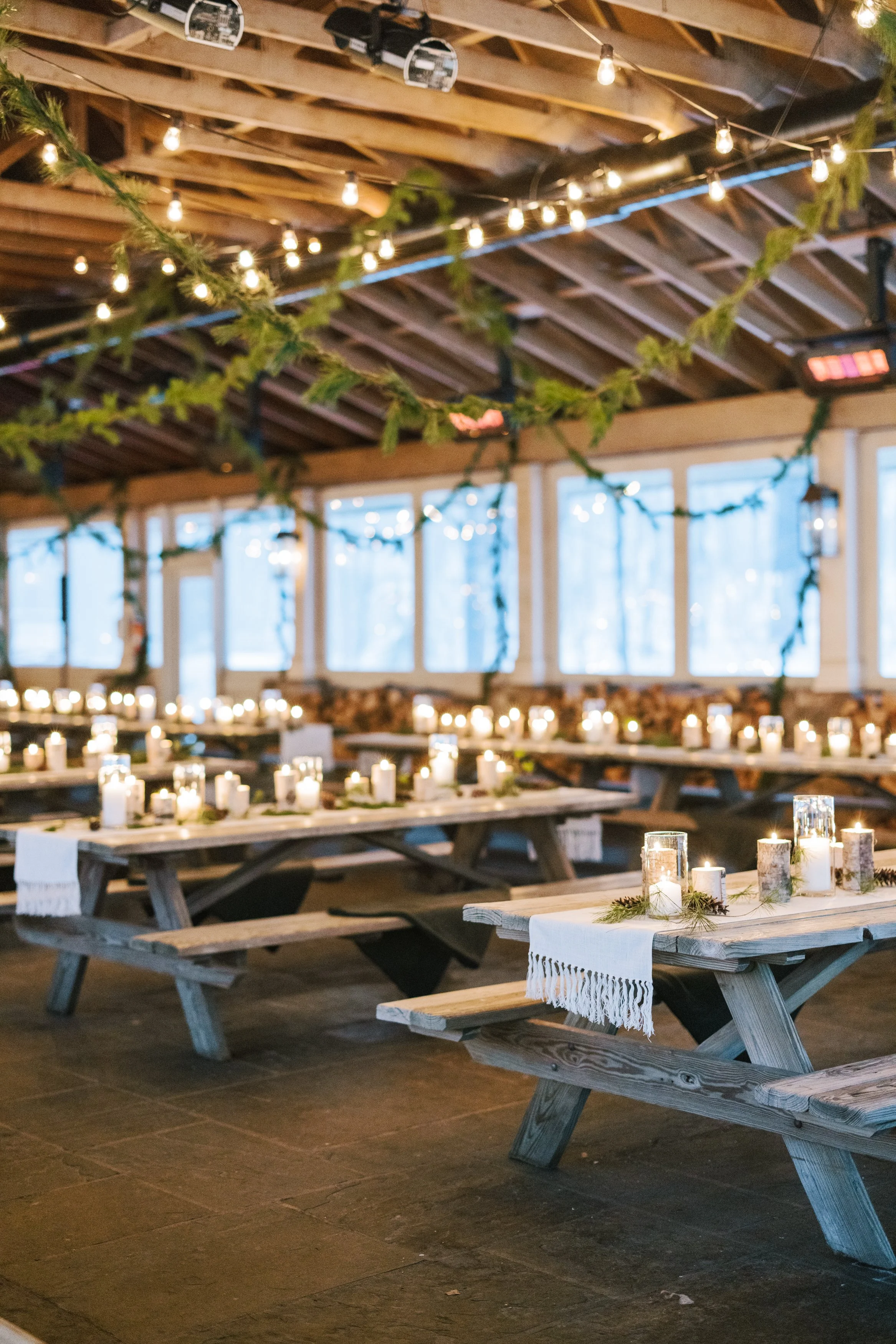 Indoor rustic setting with wooden picnic tables decorated with candles in glass holders and greenery, string lights hanging from the ceiling, and large windows in the background.