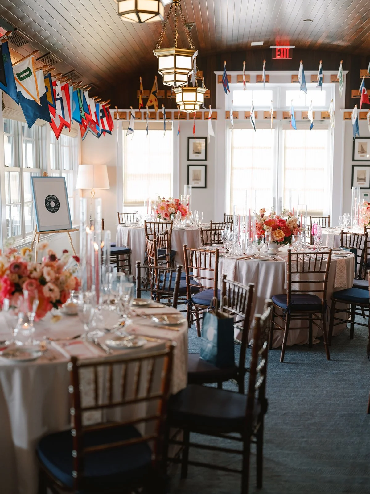 Indoor event space decorated with flags hanging from the ceiling, round tables with white tablecloths and floral centerpieces, set with glassware and silverware, large windows allowing natural light, and framed pictures on the walls.