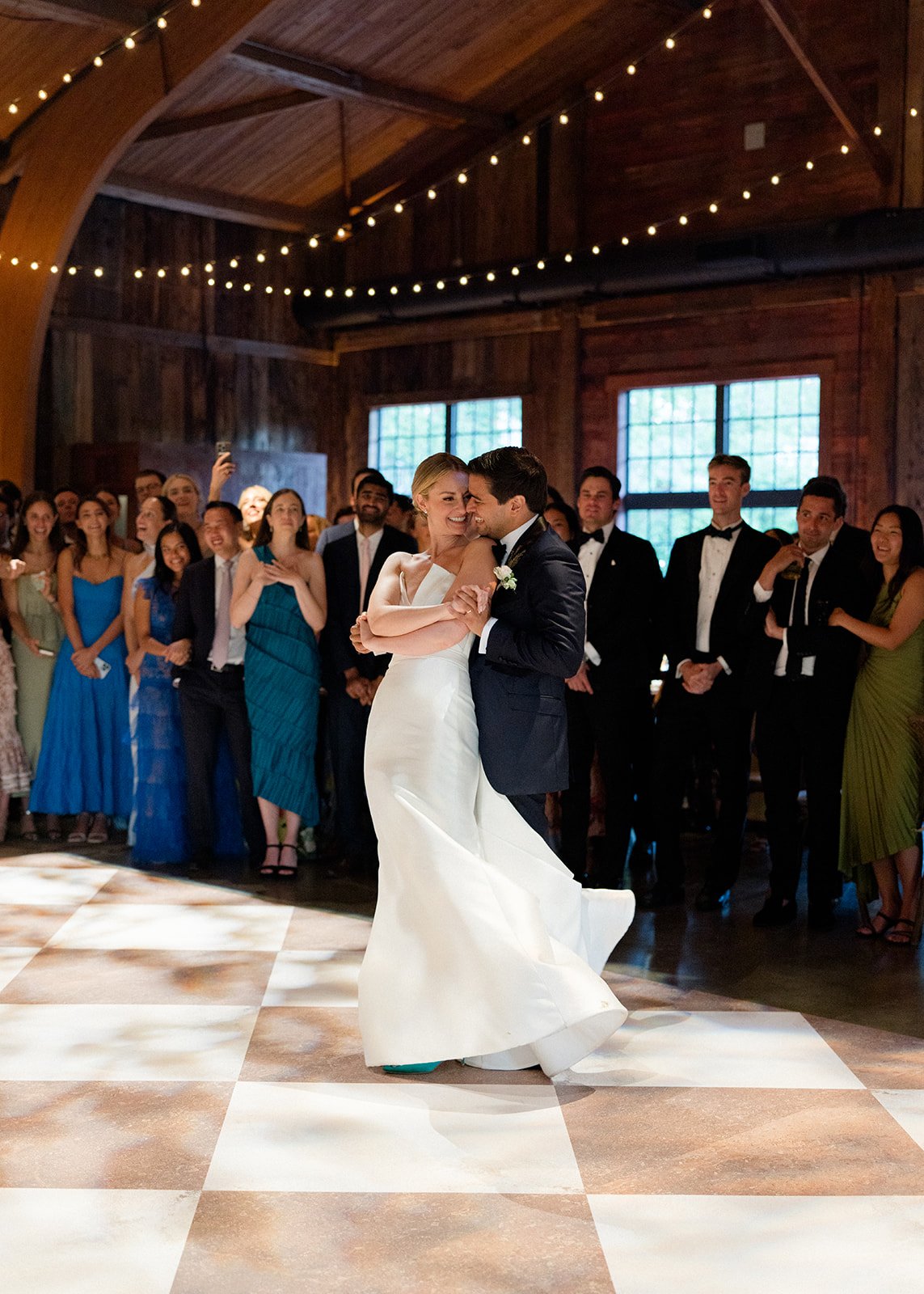 A bride and groom dancing at their wedding reception, with guests watching and smiling in a rustic wooden venue decorated with string lights.