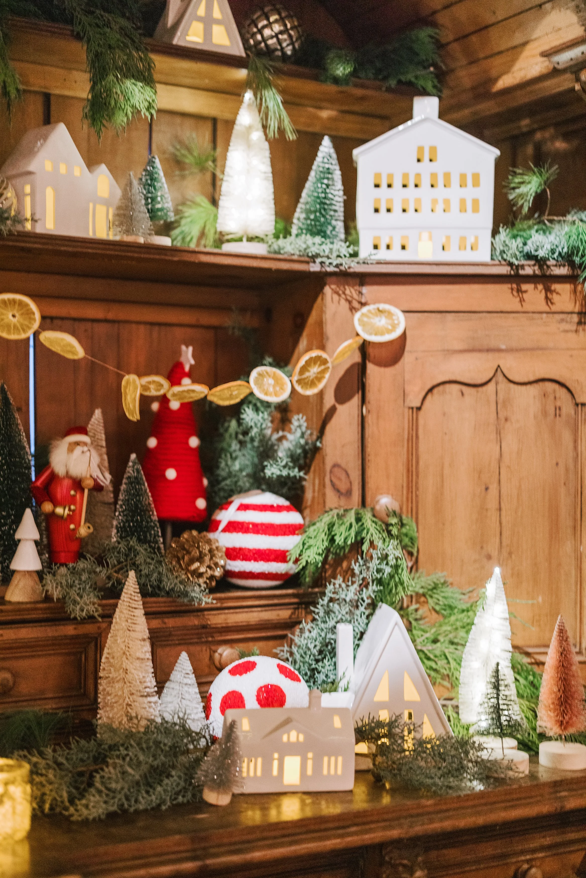 A festive holiday display with miniature illuminated white house decorations, small decorated Christmas trees, a plush Santa figure, a red and white striped ball, and a garland of dried orange slices, all arranged on a wooden surface with greenery an