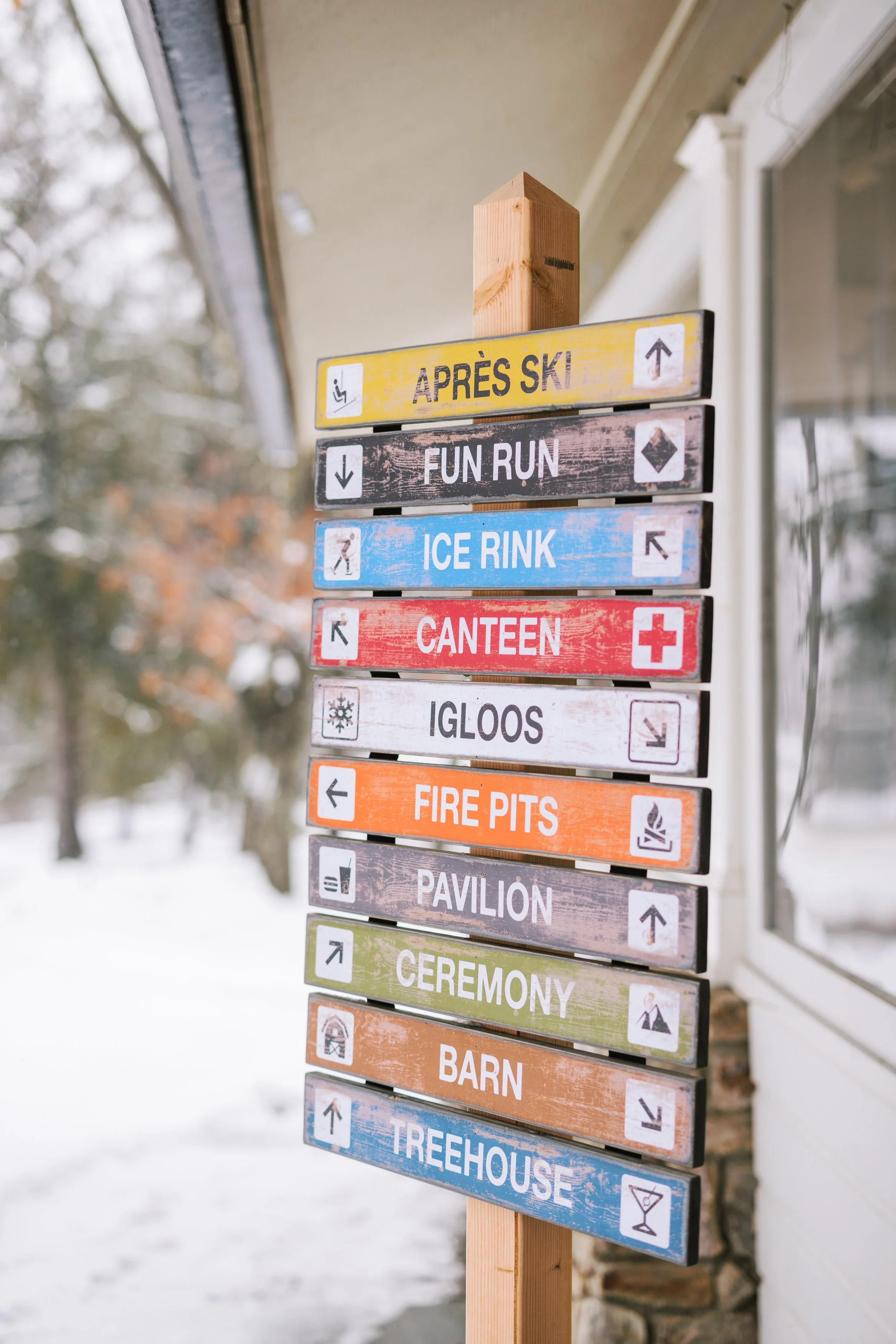 Colorful wooden signpost outdoors with directions to Après Ski, Fun Run, Ice Rink, Canteen, Igloos, Fire Pits, Pavilion, Ceremony, Barn, and Treehouse, on a snowy day.