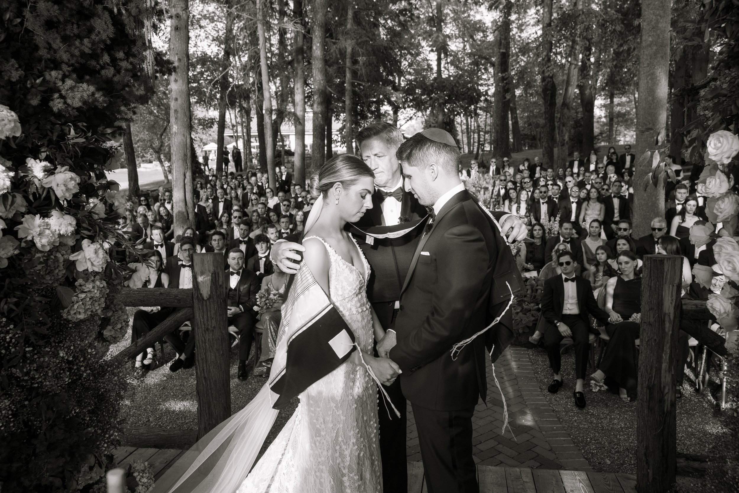 A wedding ceremony outdoors with a priest, bride, and groom standing close with heads bowed, holding hands, surrounded by a large crowd of guests seated on benches, and trees in the background.