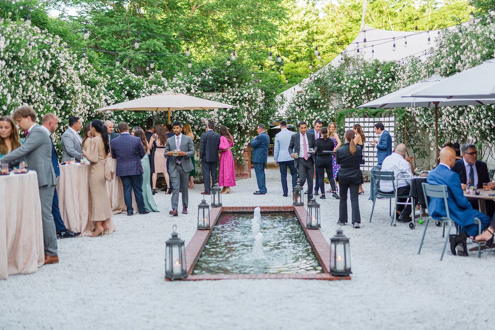 People attending an outdoor wedding reception or event, standing and sitting around tables, with string lights overhead, trees and white flowers in the background, a small water fountain in the center, and umbrellas providing shade.