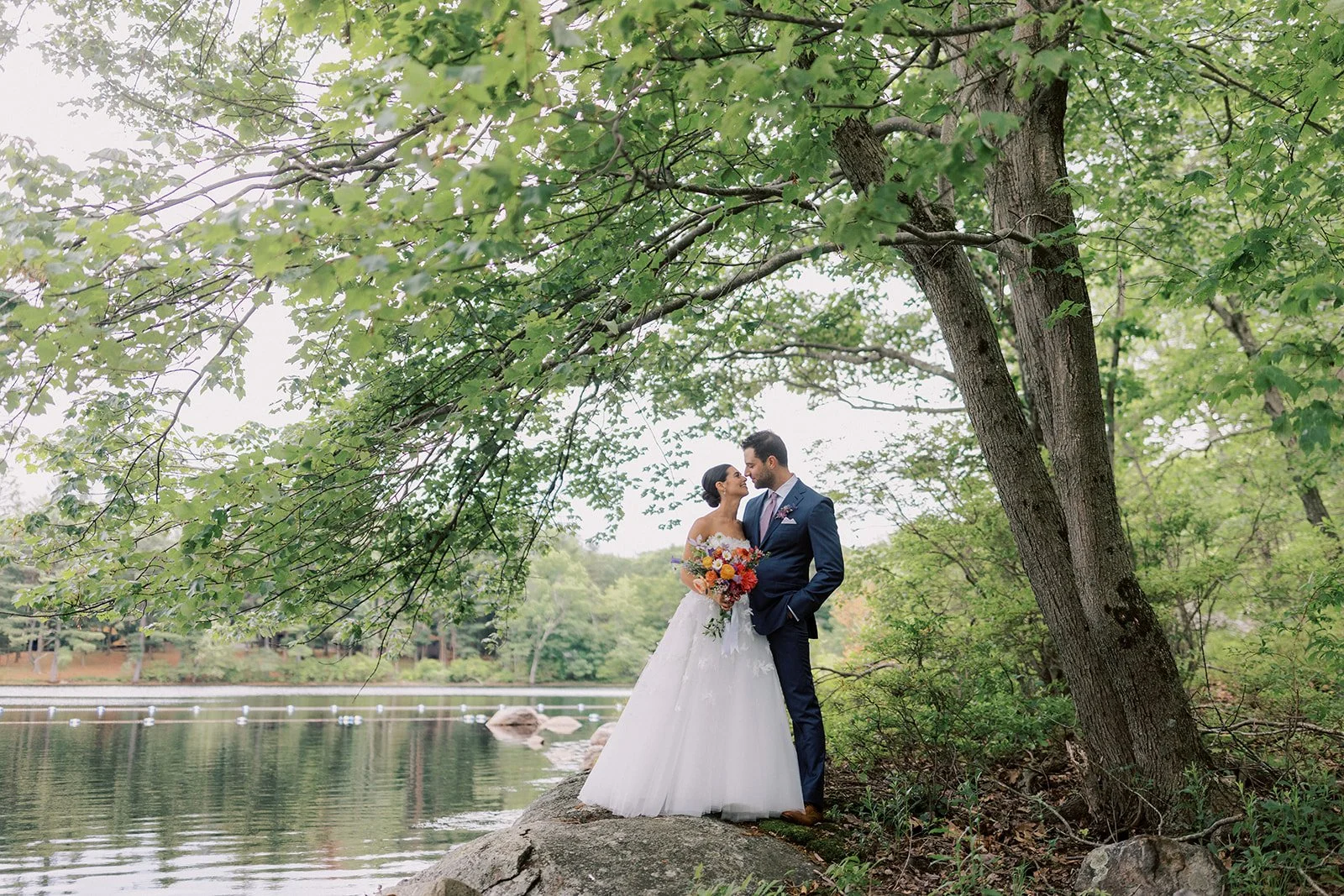 A bride and groom stand on a rock near a lake in a green forest, gazing at each other, with the bride holding a colorful bouquet.