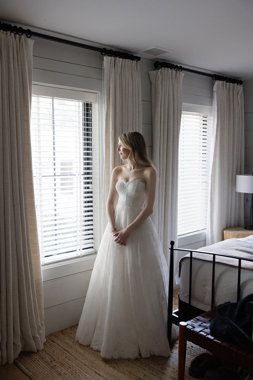 A bride wearing a strapless white wedding gown standing by a window with white blinds and beige curtains in a softly lit room.
