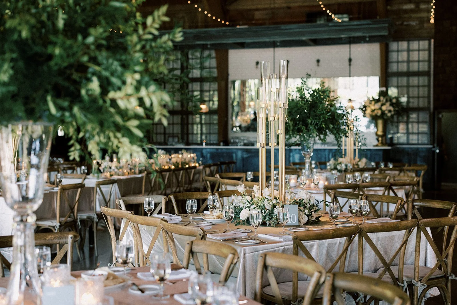 Elegant banquet hall decorated for a wedding or special event with floral centerpieces, candles, and glassware on round tables with wooden chairs, warm lighting, and greenery accents.