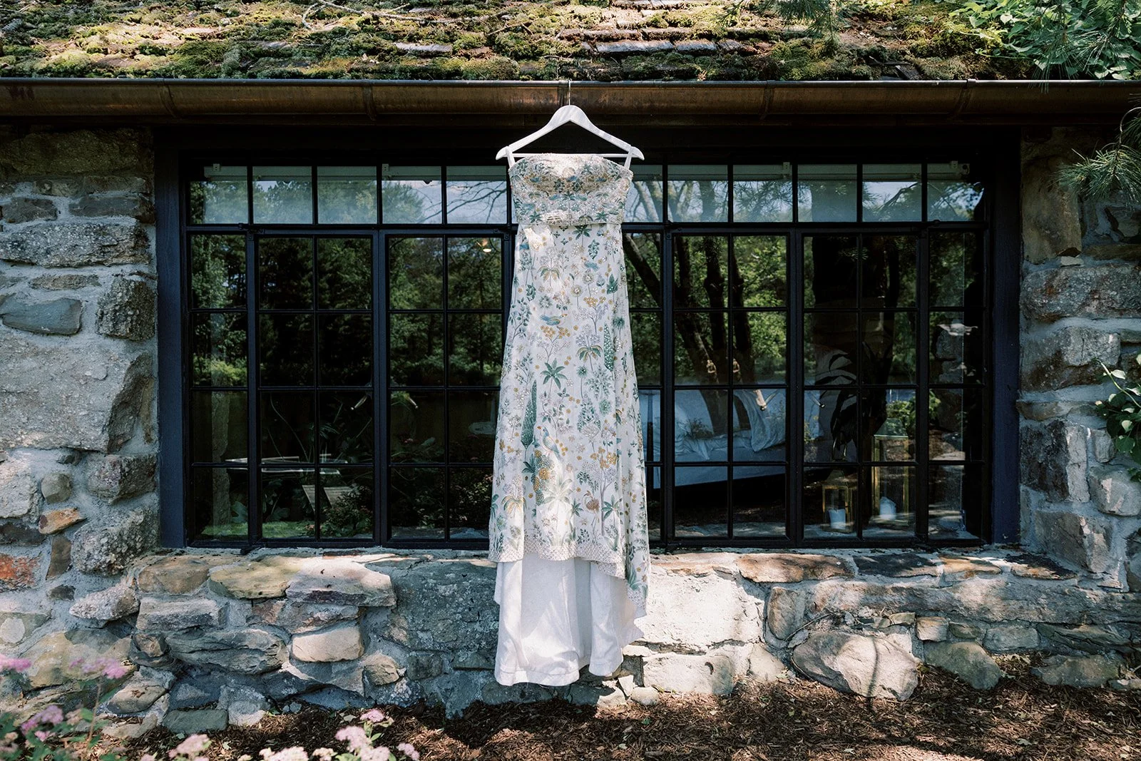 A floral dress hangs on a white hanger in front of a black-framed window on a stone wall. The dress is outdoors with trees visible through the window.